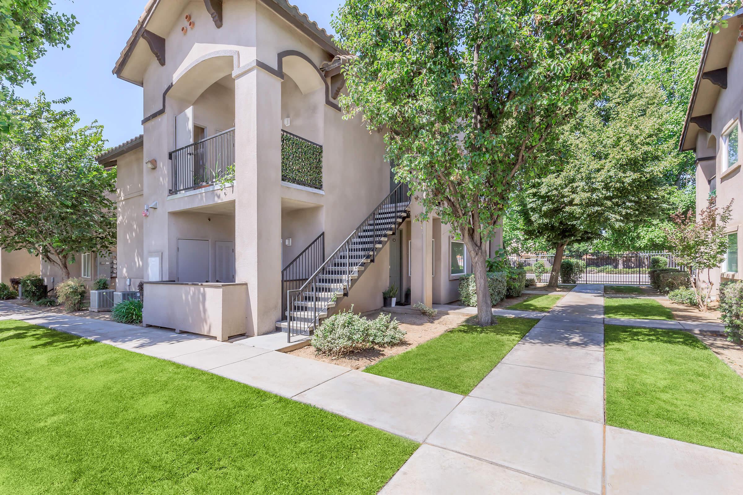 A well-maintained apartment complex with a beige building featuring a staircase and balcony. Lush green lawns and trees surround the pathways, creating a pleasant outdoor environment. The sunny day adds to the inviting atmosphere of the residential area.