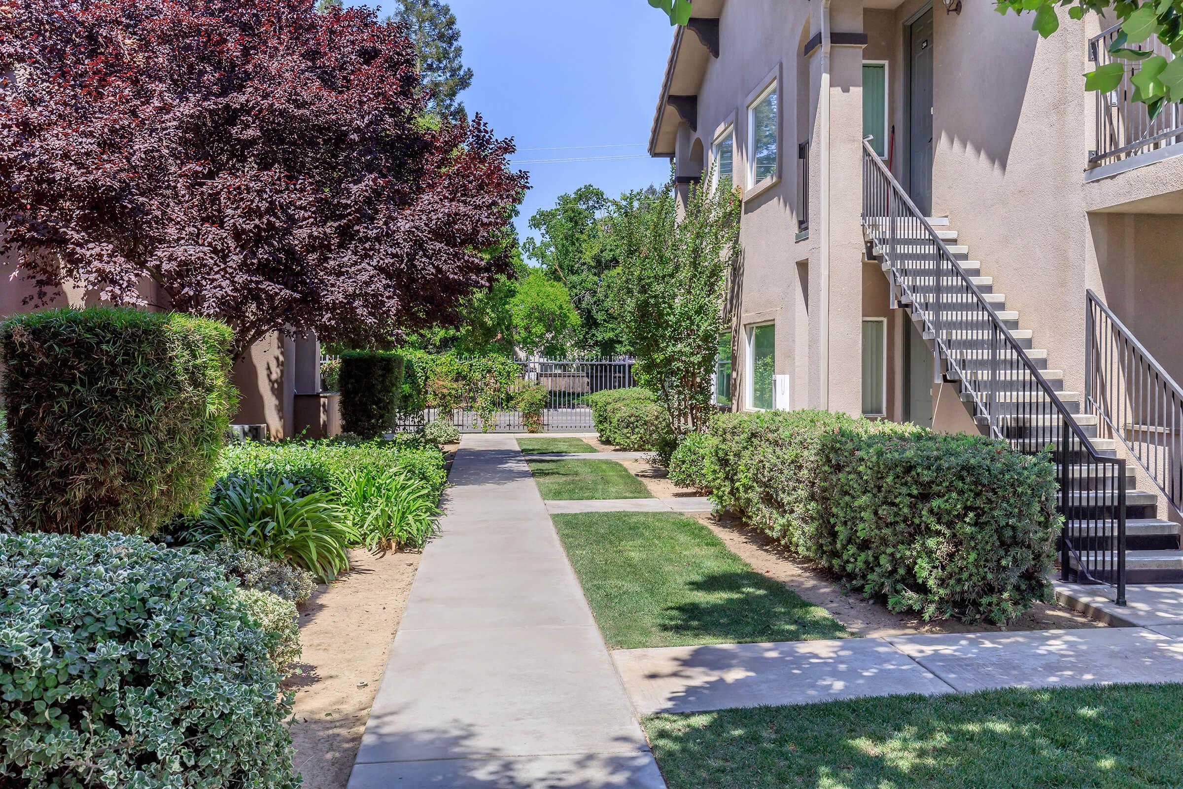 A sunny pathway runs between well-maintained hedges and greenery, leading towards an apartment complex. On one side, there are flowering bushes and a small tree, while the other features a staircase leading to upper-floor apartments. The scene is bright, with clear blue skies in the background.