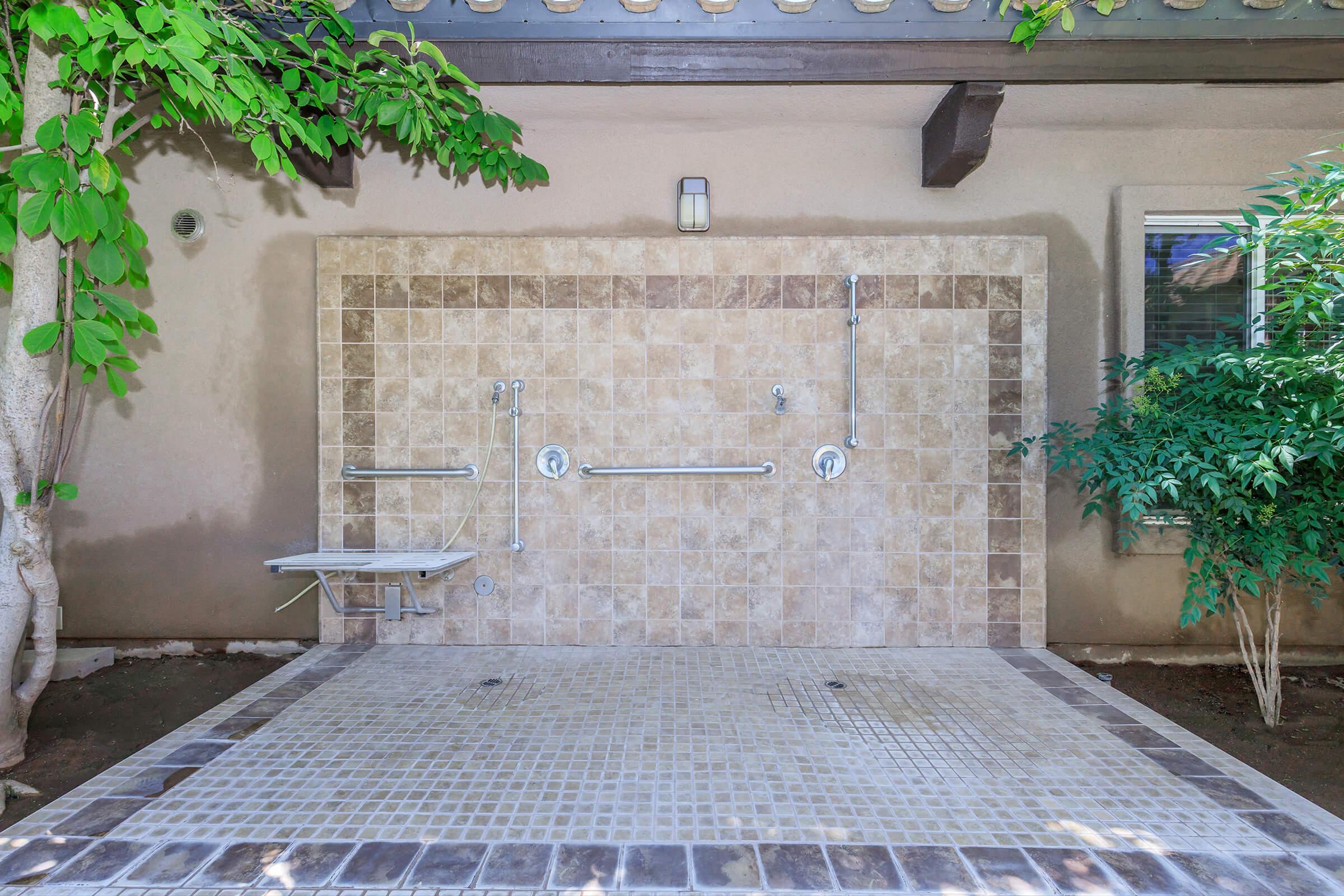 An outdoor shower area featuring a tiled wall with two showerheads, grab bars, and a small bench. Surrounding the area are lush green plants and a beige wall that complements the tiled floor beneath the shower. Natural light filters in, creating a serene ambiance.