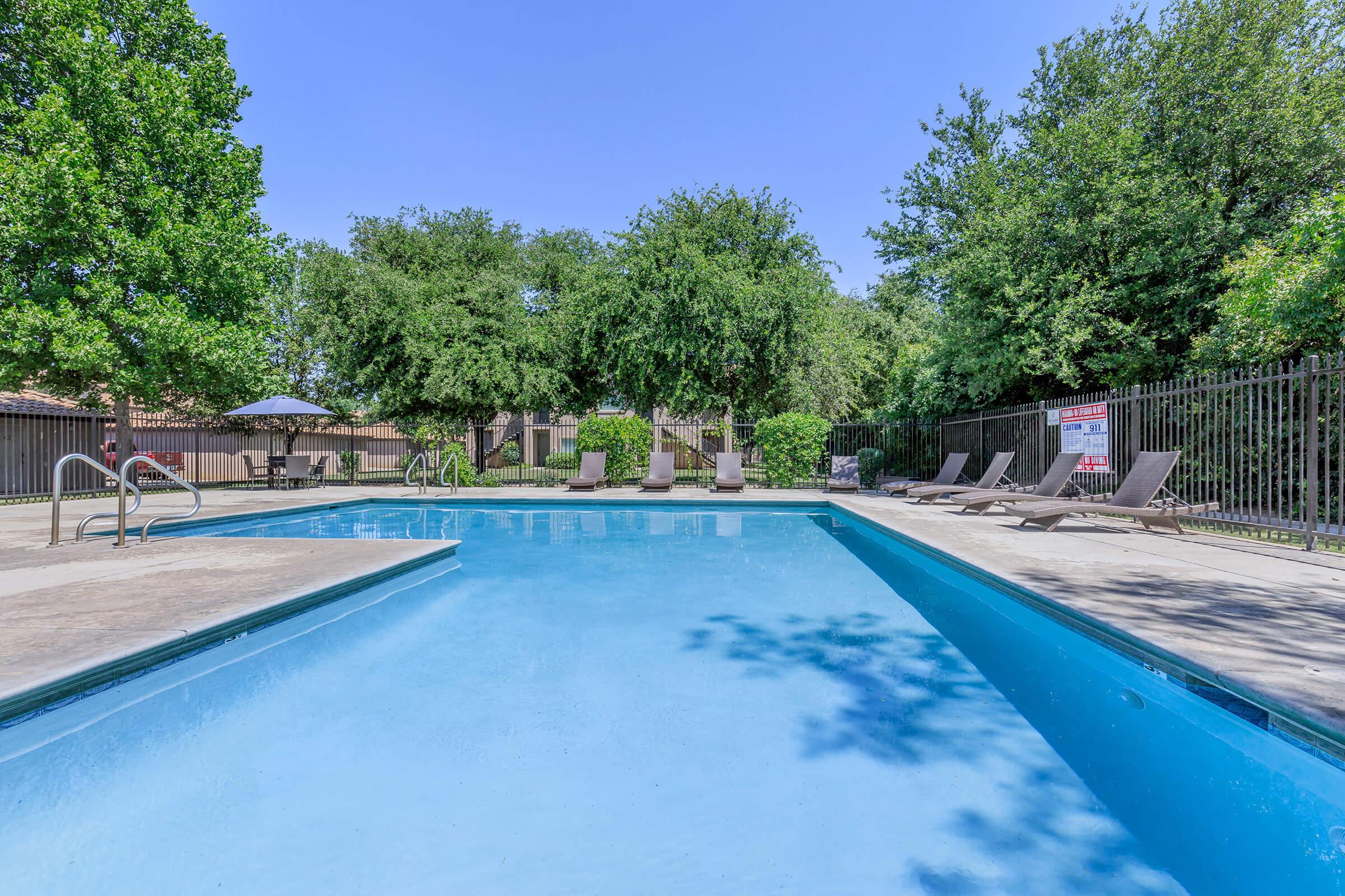 A serene outdoor swimming pool surrounded by lush greenery. The clear blue water reflects the sunlight, with lounge chairs arranged around the pool area. Shade is provided by trees and an umbrella, creating a relaxing atmosphere for lounging and enjoying the outdoors.