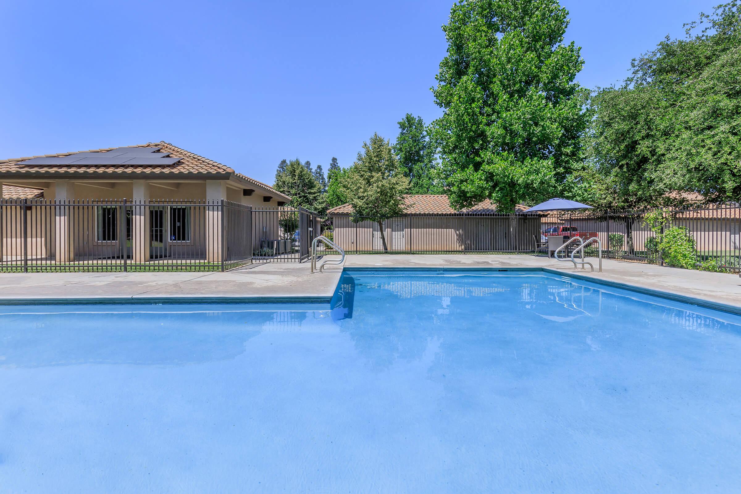 A clear swimming pool surrounded by a paved deck, with a fence in the background. Lush trees provide shade, and nearby buildings feature a tile roof. The sky is bright blue, creating a sunny and inviting atmosphere. Steps lead into the pool, perfect for lounging and relaxation.