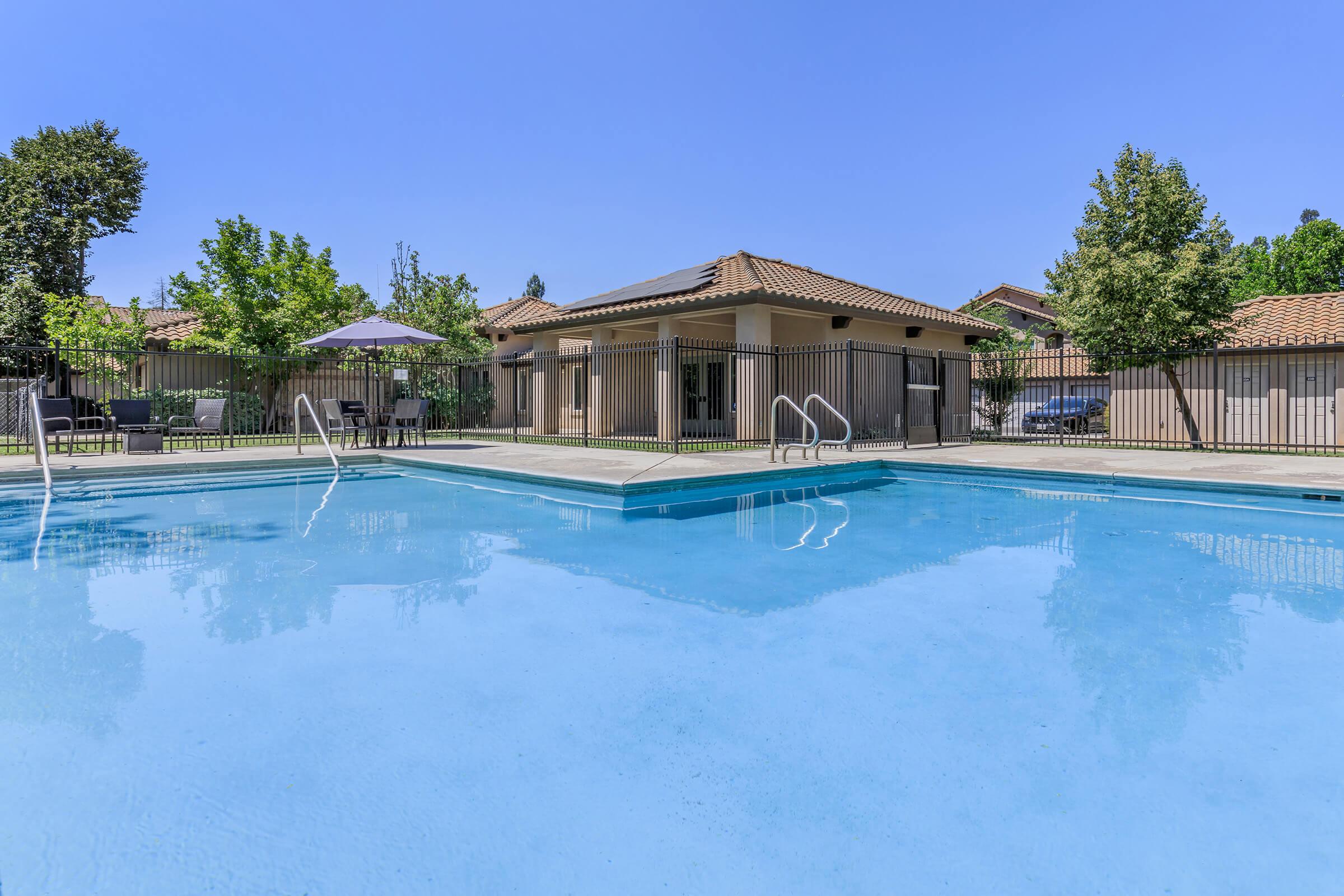 A clear blue swimming pool with diving steps in the foreground, surrounded by a wooden fence. In the background, a patio area with tables and umbrellas, and a building with a tiled roof. Lush green trees provide shade, under a bright blue sky.