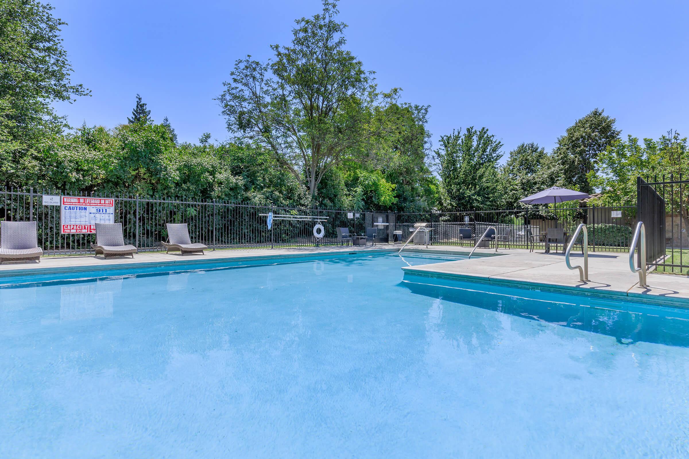 A clear swimming pool surrounded by lounge chairs and a poolside area. Lush greenery and trees provide a backdrop, with a sunny blue sky overhead. A lifebuoy is mounted along the pool's edge, and a large umbrella offers shade. The scene is inviting and tranquil, ideal for relaxation on a warm day.
