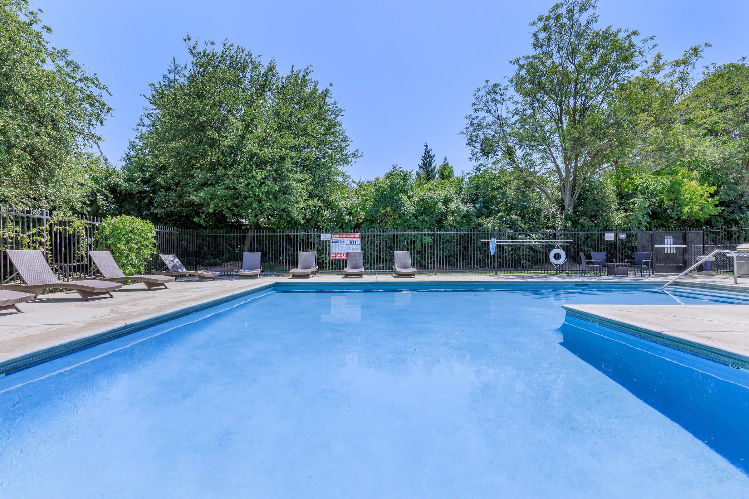 A clear blue swimming pool surrounded by lounge chairs, with trees and greenery in the background. A poolside fence encloses the area, and a sign is visible on the fence. The sky is sunny and blue, creating a calm and inviting atmosphere.