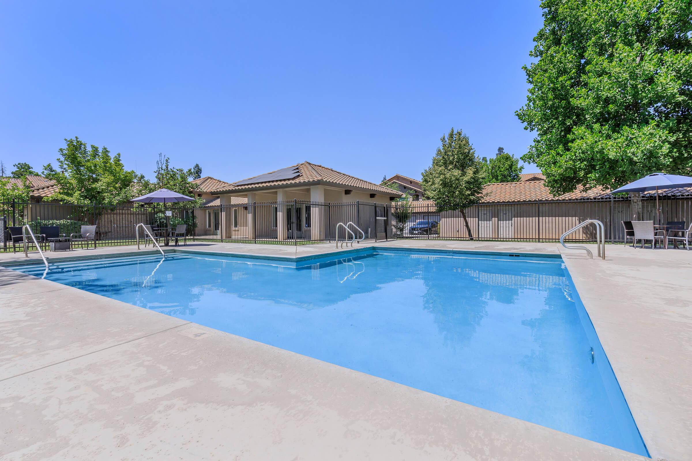 A clear blue swimming pool surrounded by a concrete deck, patio furniture, and umbrellas. Lush green trees and a building with a tiled roof are in the background, under a bright blue sky. The setting appears calm and inviting, ideal for relaxation and leisure.