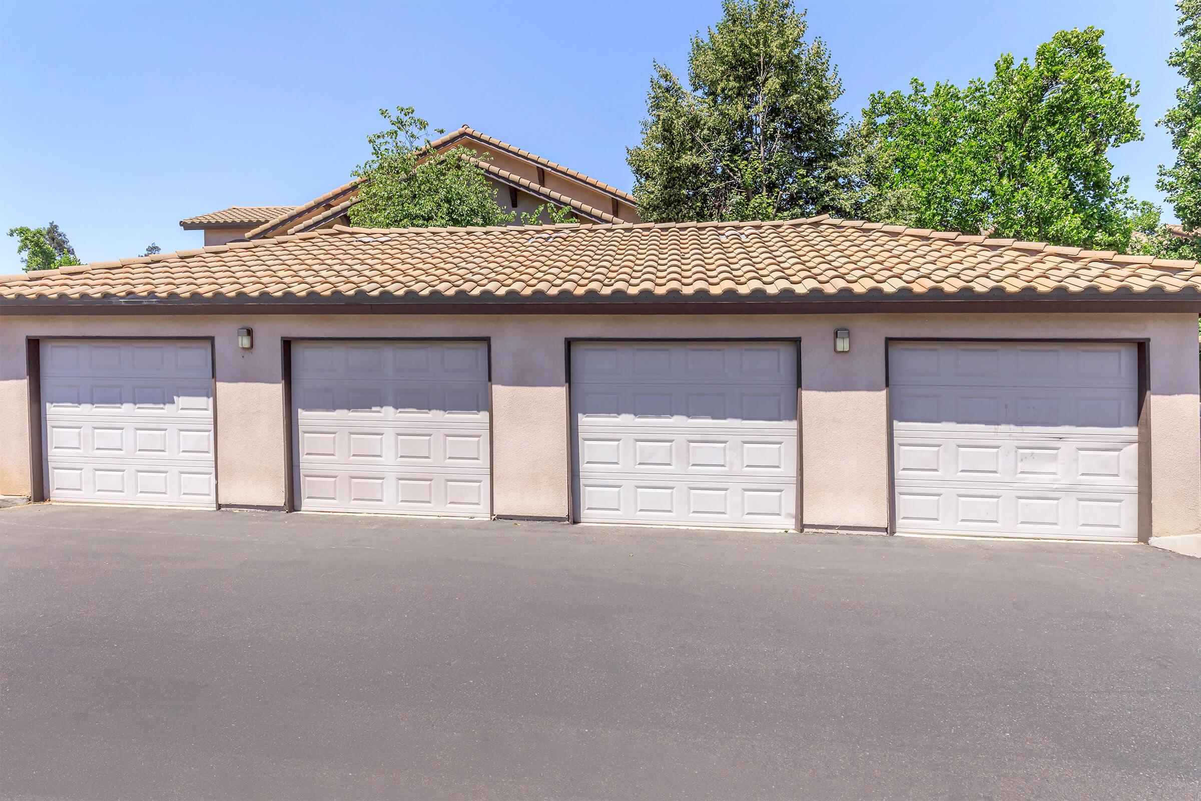 A row of three closed garage doors with white paneling set against a light-colored stucco wall. The garages are accompanied by a paved driveway and a house with a tiled roof in the background, surrounded by green trees. The sky is clear and blue.
