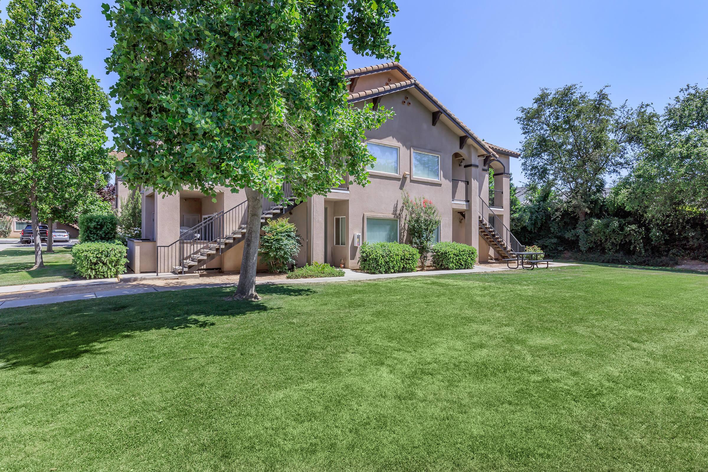 A two-story residential building with a beige exterior, surrounded by neatly trimmed grass and trees. The building features multiple windows and a staircase leading to the upper level. A small picnic area with benches is visible on the lawn, providing a pleasant outdoor space. Clear blue sky overhead.