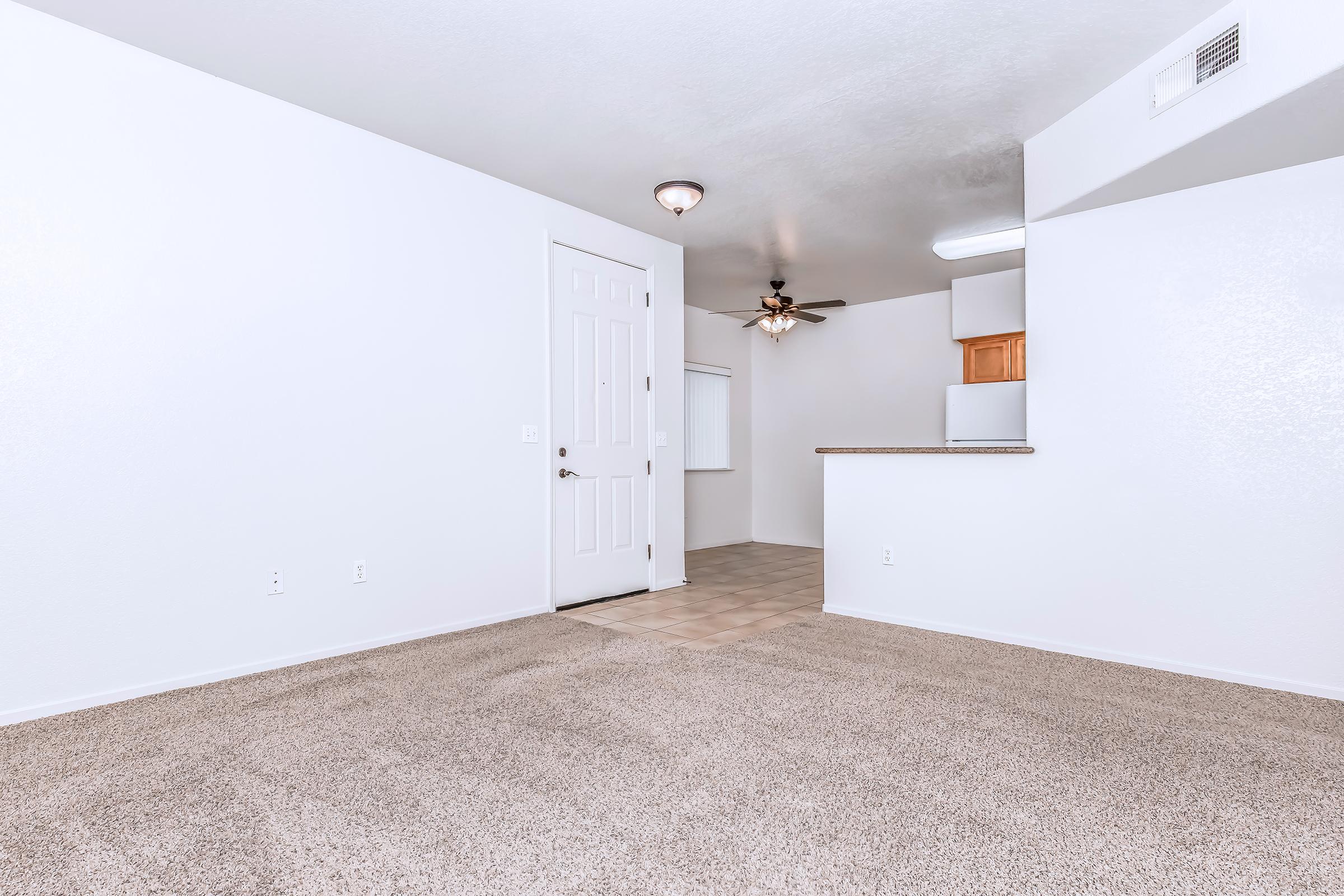 Interior of a modern apartment featuring light-colored walls and carpet flooring. An open layout includes a door leading outside, a ceiling fan, and a view into a kitchen area with wooden cabinets. The space is bright and uncluttered, creating a welcoming atmosphere.