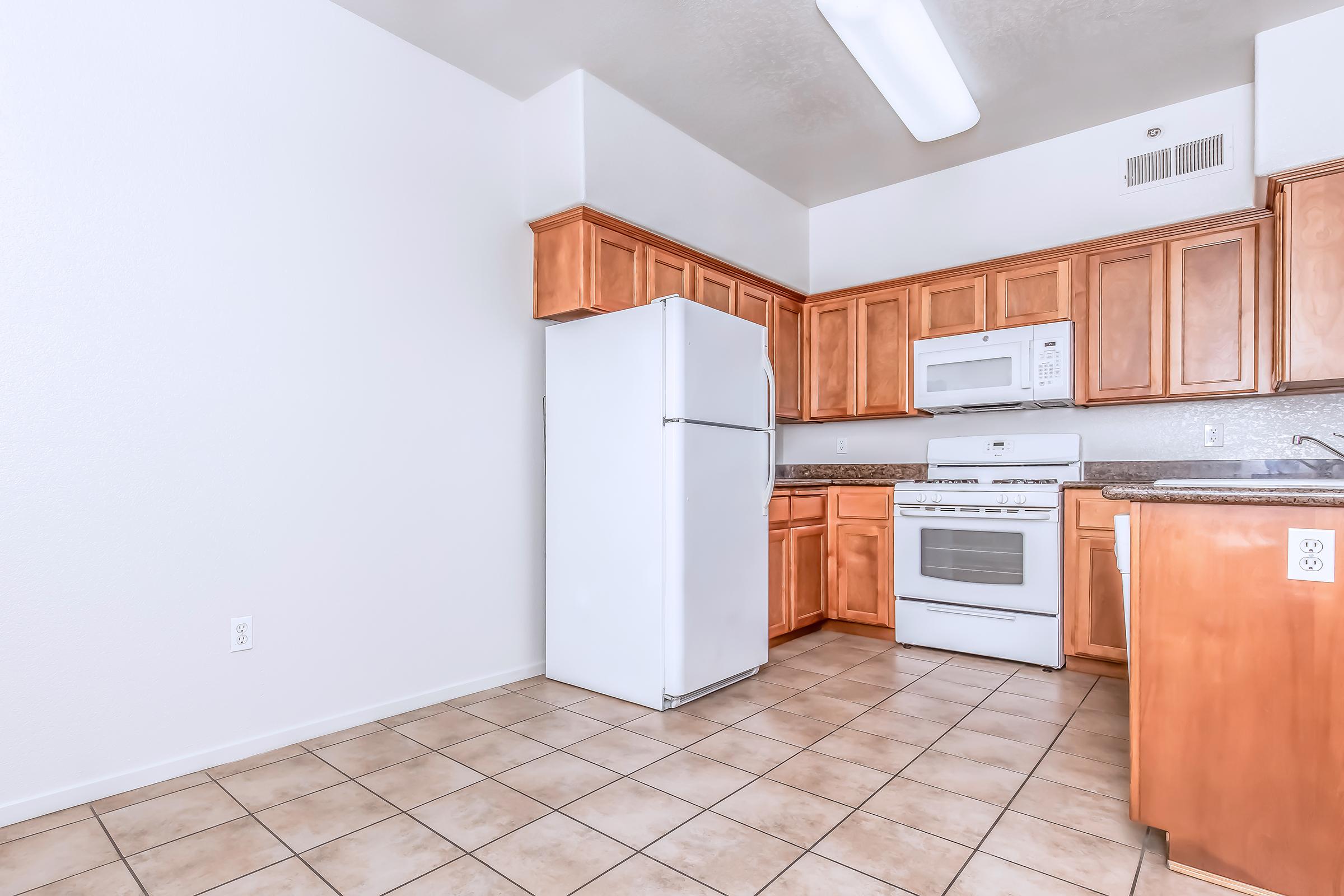 A modern kitchen featuring wooden cabinetry, a white refrigerator, and a white stove with an oven. The countertops are granite, and the flooring consists of beige tiles. A bright overhead light illuminates the space, creating a clean and inviting atmosphere.