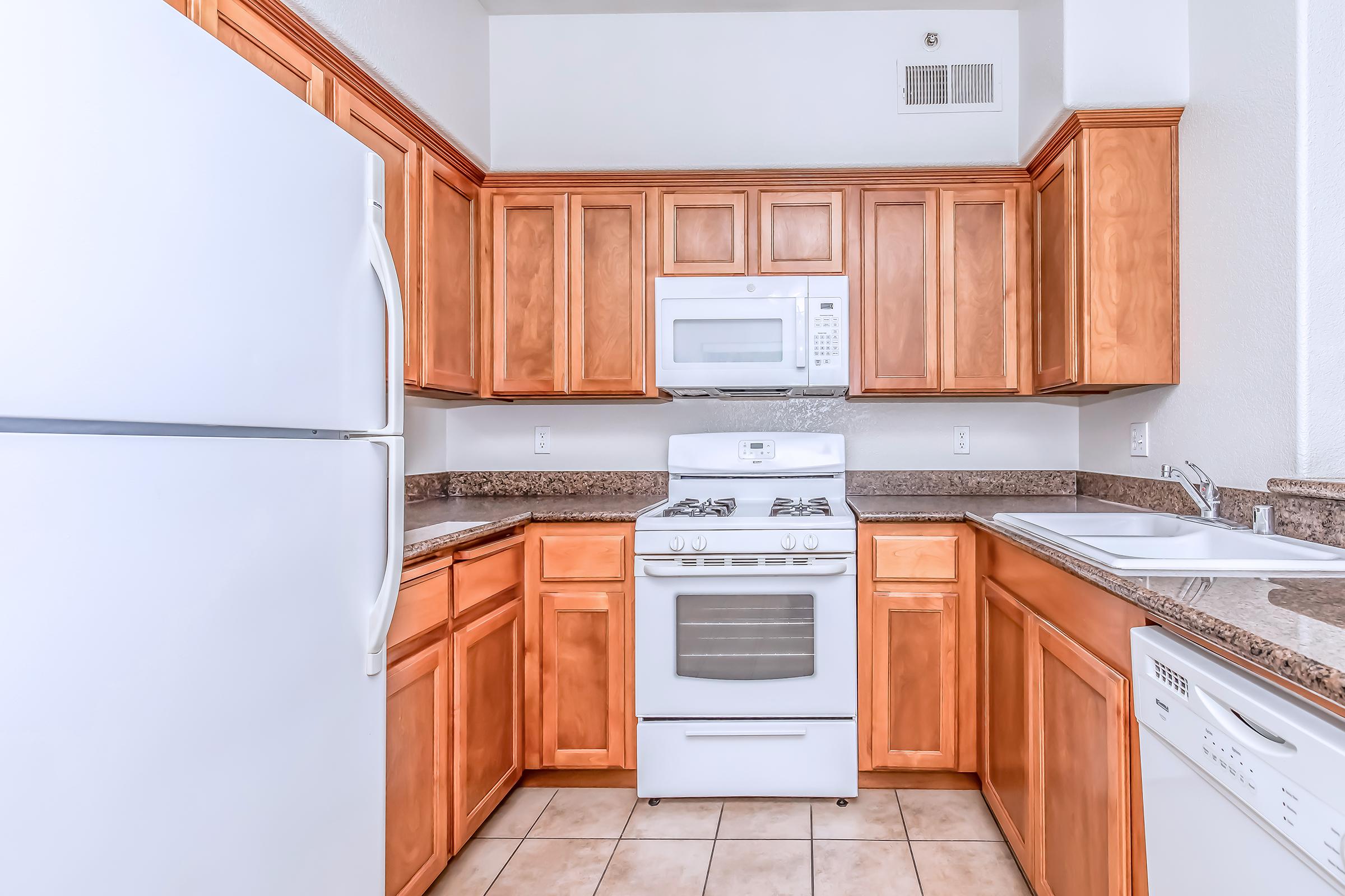 A modern kitchen featuring wooden cabinets, a white refrigerator, a white gas stove with an oven, and a microwave above it. The countertop is made of granite, and there's a sink with a faucet. The tile flooring complements the warm tones of the cabinetry.