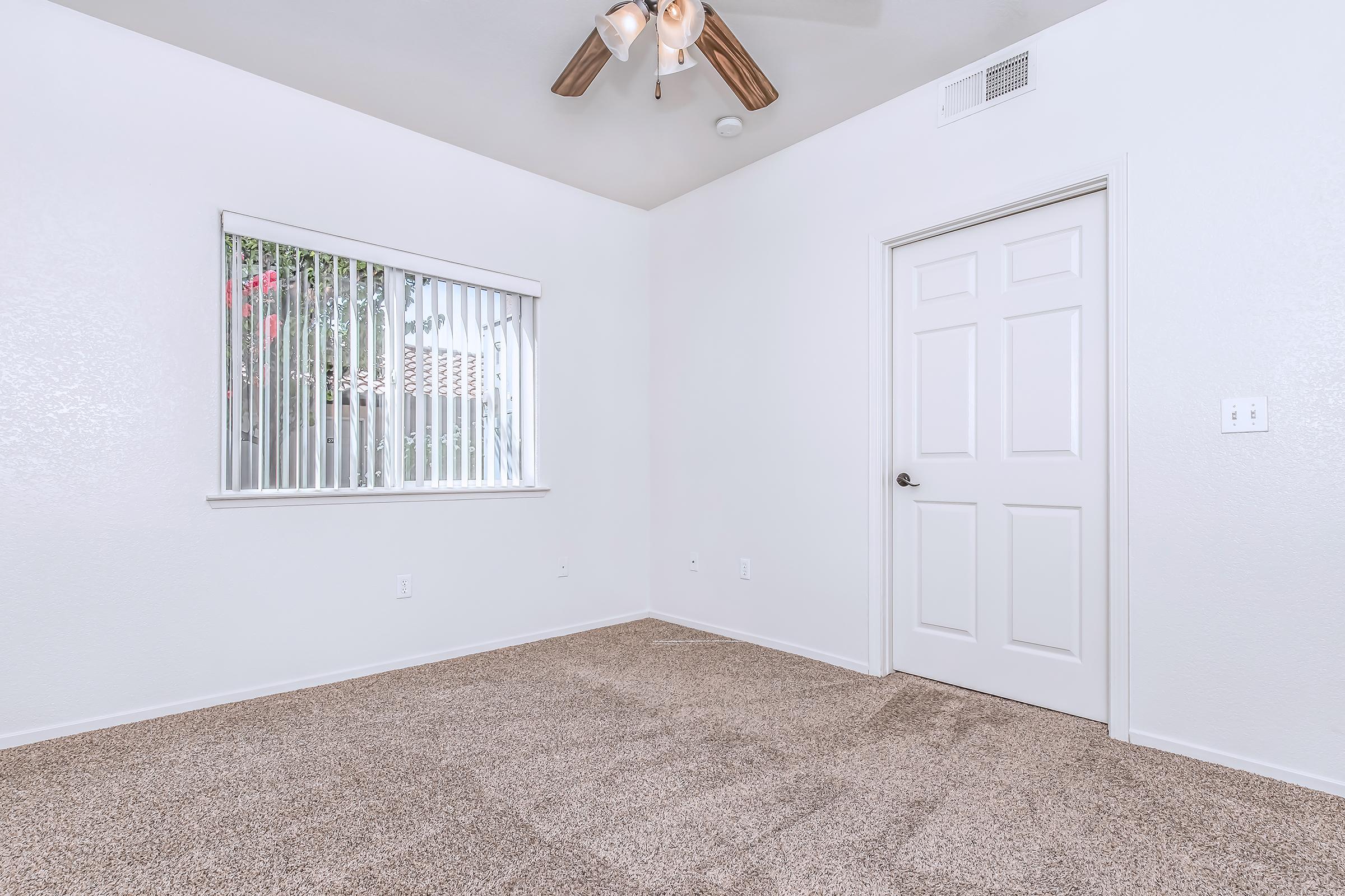 A clean, empty room with beige carpeting and white walls. A ceiling fan with wooden blades is installed, and there is a window with vertical blinds letting in natural light. A closed door is visible on one wall, and the overall atmosphere is bright and minimalist.