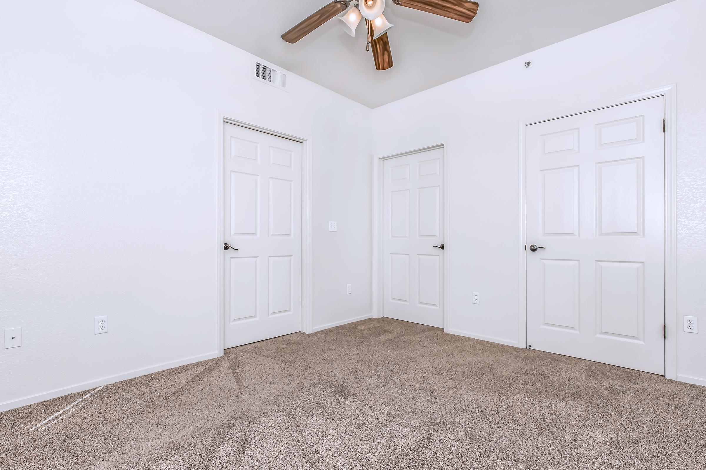 A spacious room with light-colored walls and beige carpeting. It features three white doors and a ceiling fan with wooden blades. The room is empty, providing a clean and simple aesthetic, ideal for various uses. Natural light may enhance the open feel of the space.