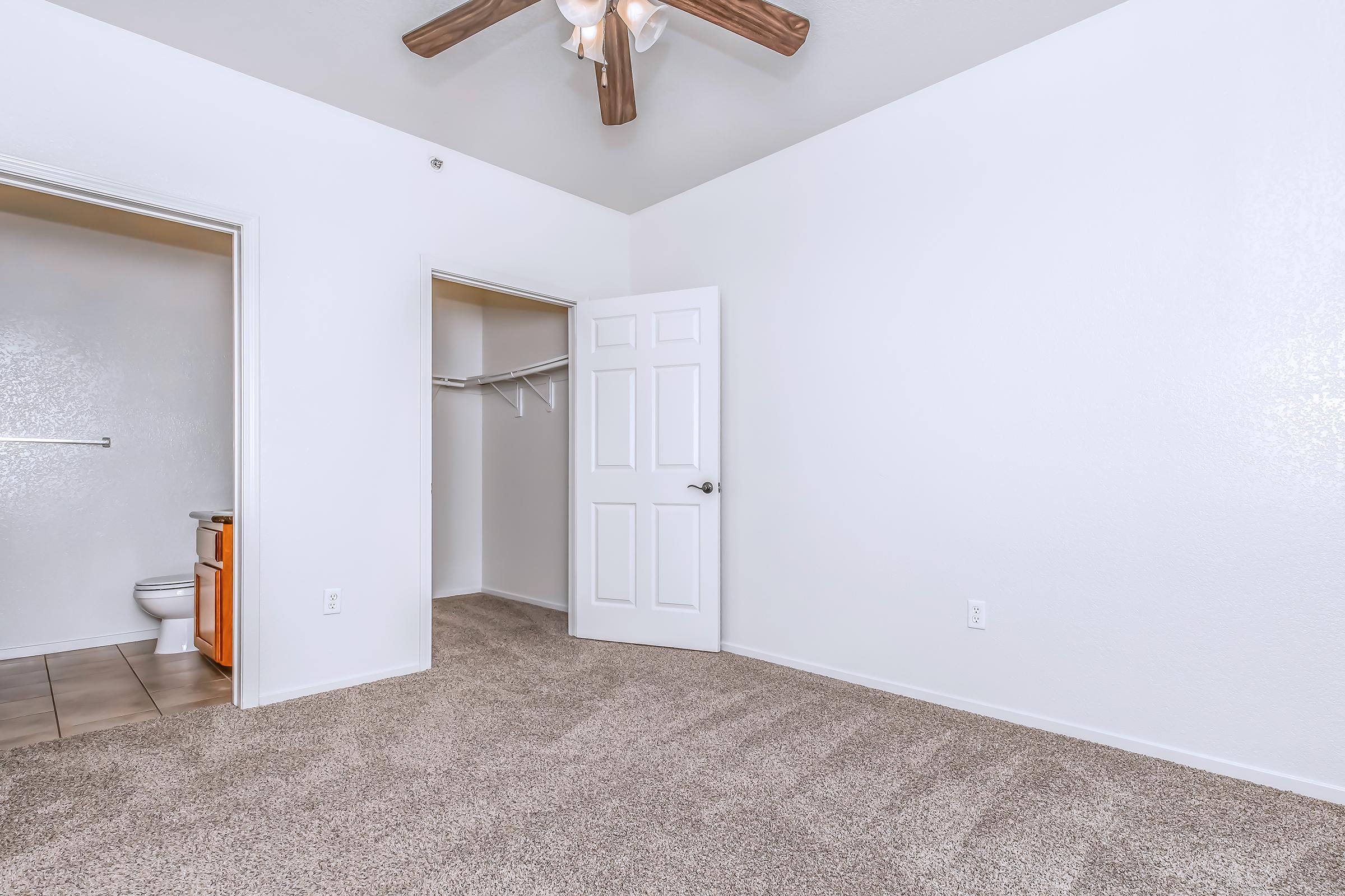 A spacious, well-lit room featuring light beige carpet and white walls. A ceiling fan is installed. On the left, there's an open door leading to a bathroom with a toilet visible. To the right, a closet with shelving is partially open. The overall atmosphere is clean and inviting.