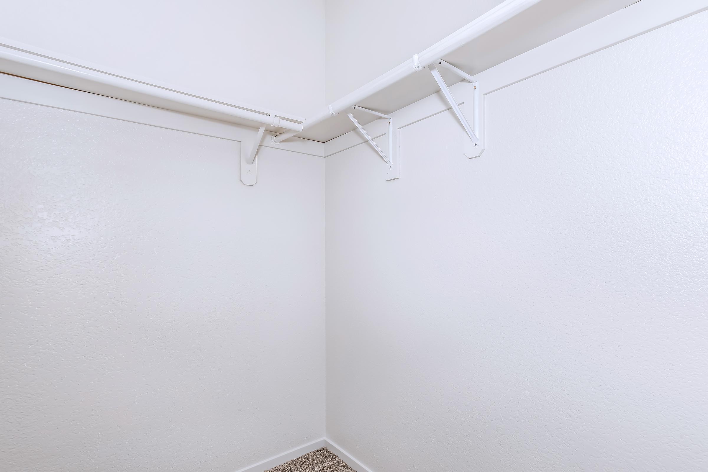 Empty closet with white walls and a carpeted floor. Two horizontal rods are mounted on the walls for hanging clothes, and there are brackets supporting the rods. The closet has a simple and clean appearance with no items stored inside.
