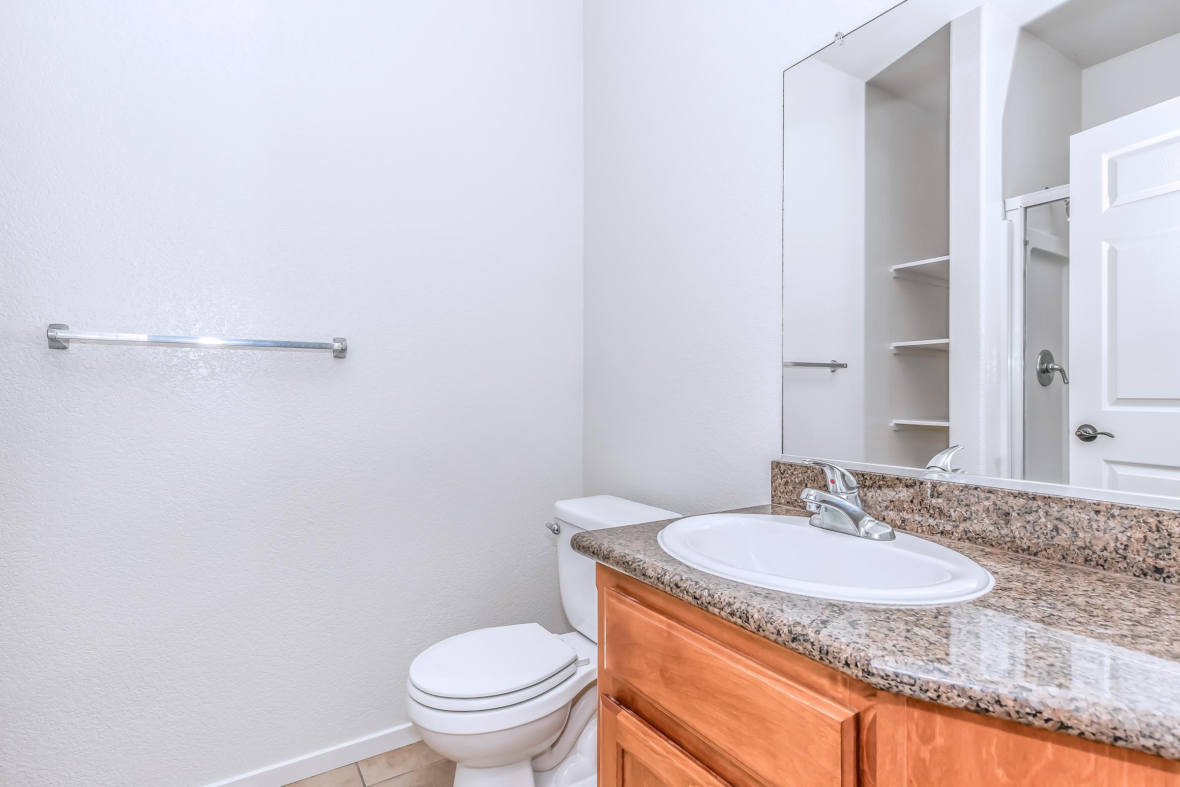 A clean and modern bathroom featuring a white toilet, a granite countertop with a sink, a large mirror, and white walls. Shelving is visible in the background, and there is a towel rack beside the sink. Natural light brightens the space.