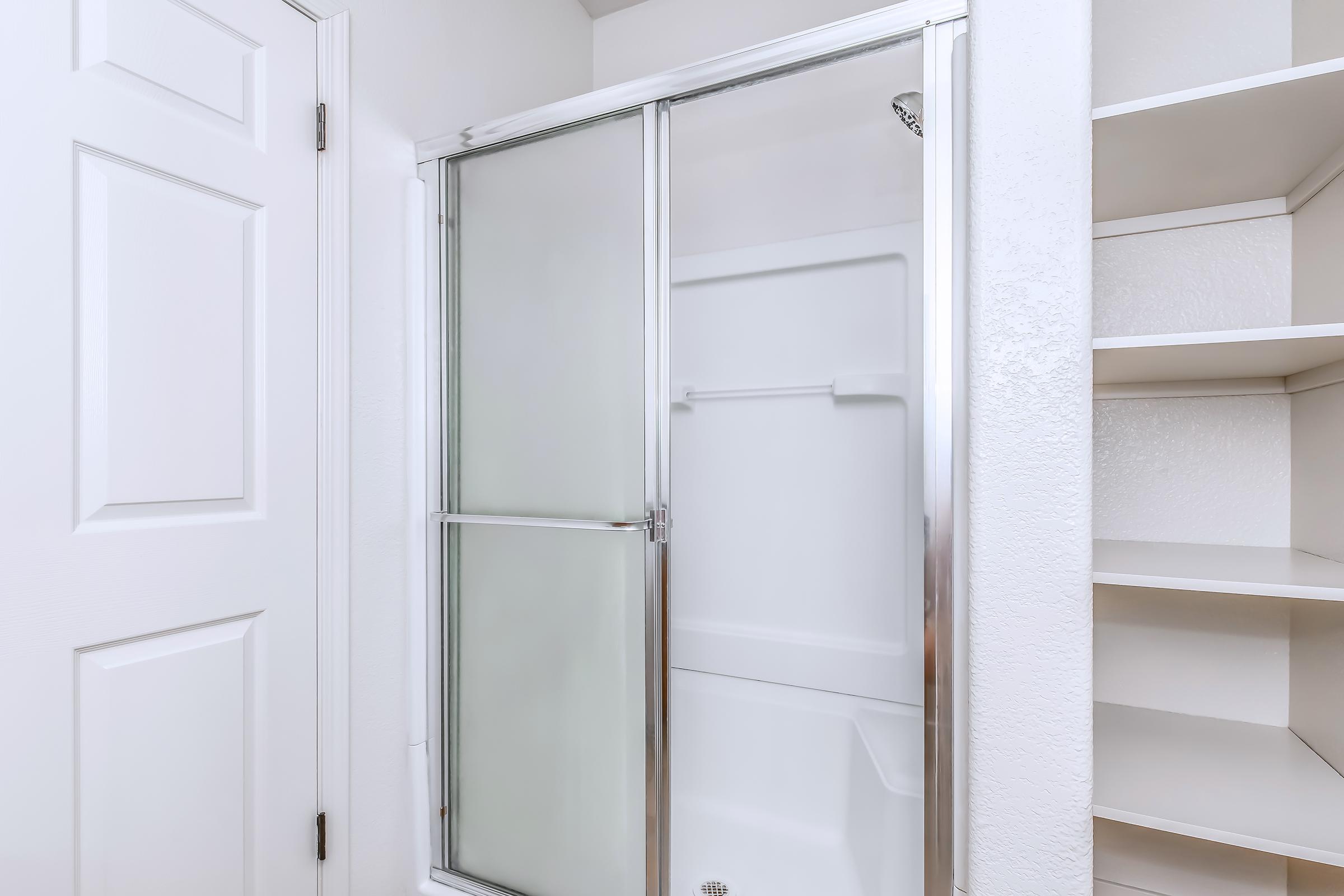 A clean, white bathroom featuring a glass shower enclosure with sliding doors. The shower is situated next to a closed door and adjacent to a built-in shelving unit. The walls are painted in a bright color, creating a fresh and modern ambiance.