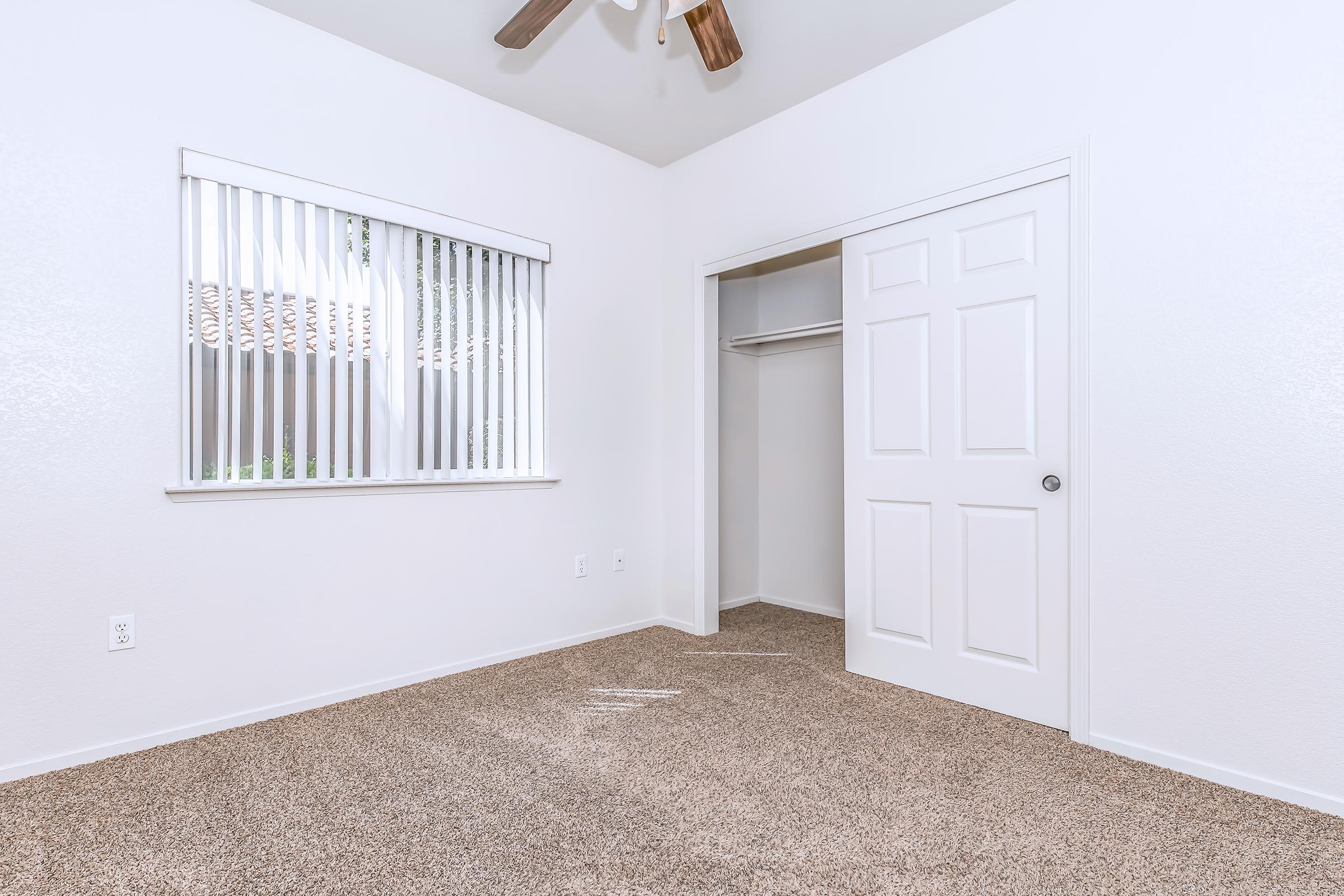 A small, clean bedroom featuring light-colored walls and beige carpet. There is a window with horizontal blinds allowing natural light in, and a closet with a white door. The room has a simple, undecorated aesthetic, making it suitable for various styles of decor.