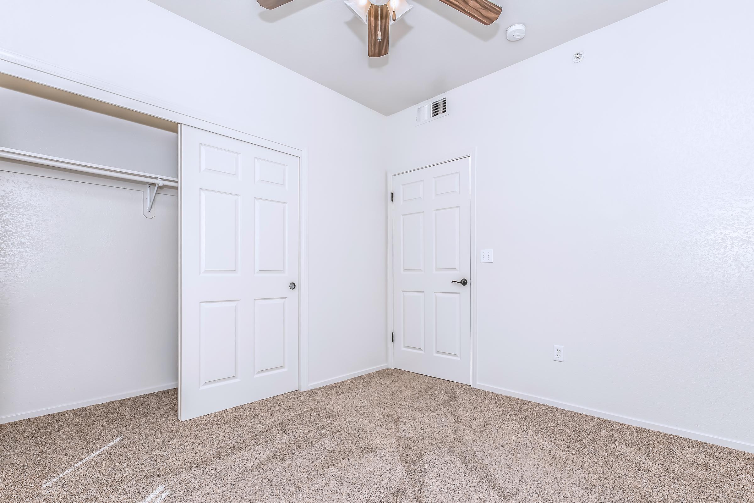 A well-lit, empty room with white walls and a light brown carpet. There are two closed doors, one leading to a closet. A ceiling fan is visible above, and the room has no furniture, emphasizing its spaciousness and cleanliness.