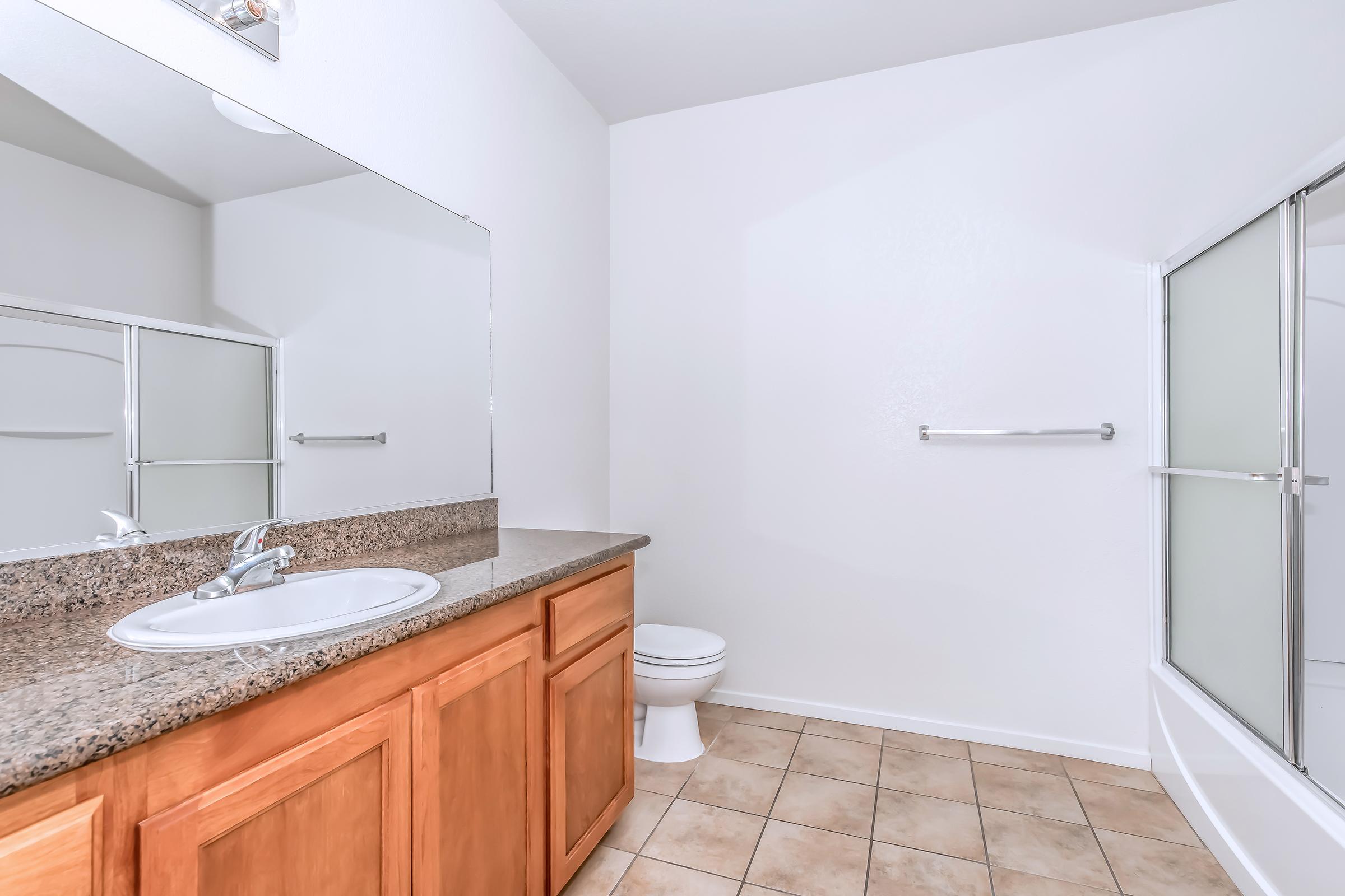 A clean and modern bathroom featuring a large mirror, a sink with a granite countertop, wooden cabinets, and a white toilet. The floor is tiled, and there's a glass-enclosed shower in the corner. The walls are painted white, contributing to a bright and airy feel.
