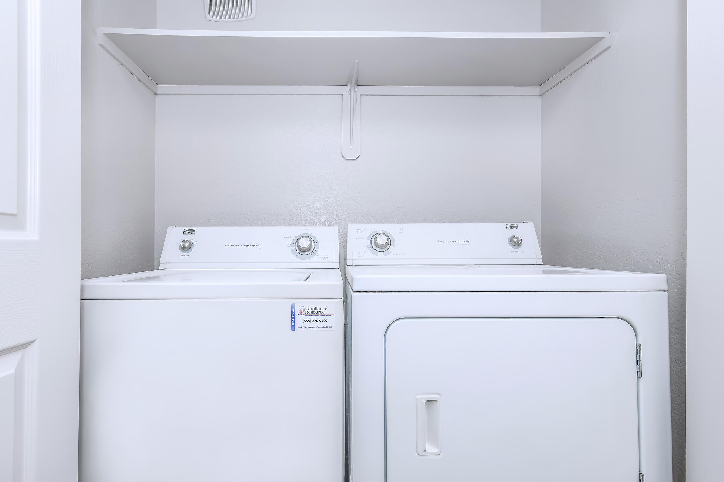 A laundry room featuring a white washing machine and a white dryer stacked next to each other. Above them is a simple shelf mounted on the wall, all set against a neutral-colored wall. The space is clean and organized, ideal for household laundry tasks.