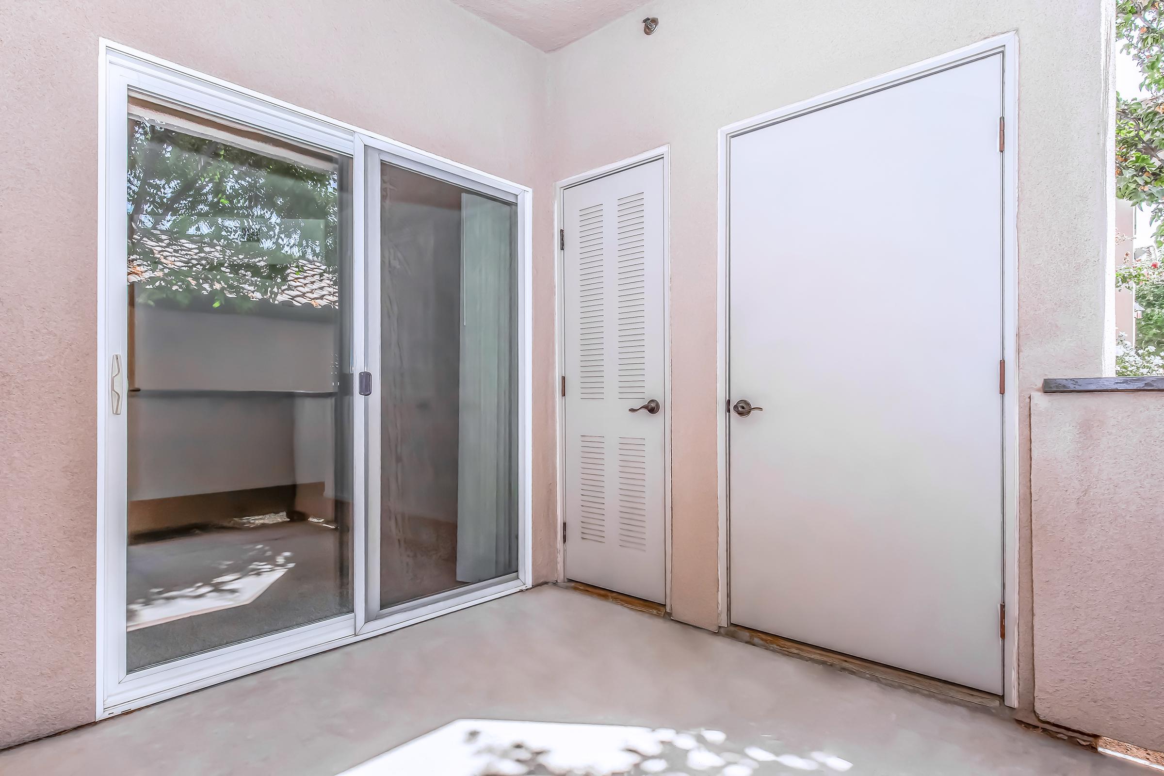 A small, well-lit patio area featuring a sliding glass door on the left leading to an outdoor space, and a closed white door on the right. The walls are painted a light color, and there are shadows cast by nearby foliage. The floor is a smooth concrete surface.