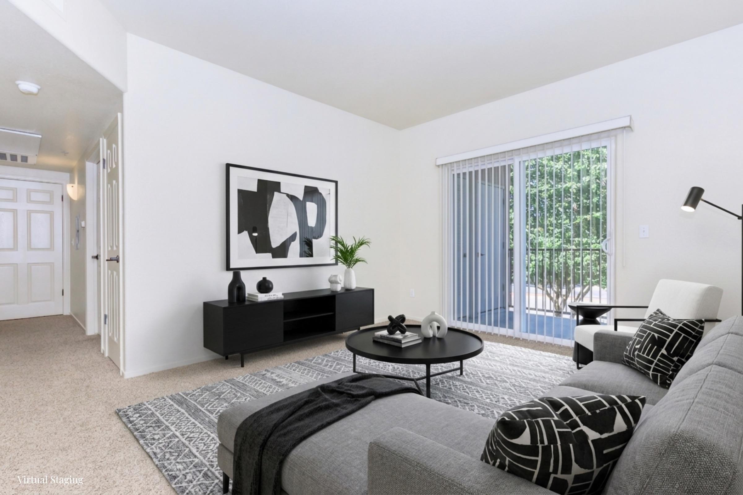 A modern living room featuring a gray sofa with patterned cushions, a round coffee table, and a minimalist black TV stand. There is a large framed abstract artwork on the wall, a potted plant on the side table, and sliding glass doors leading to a patio area. Neutral colored walls and carpet complement the decor.