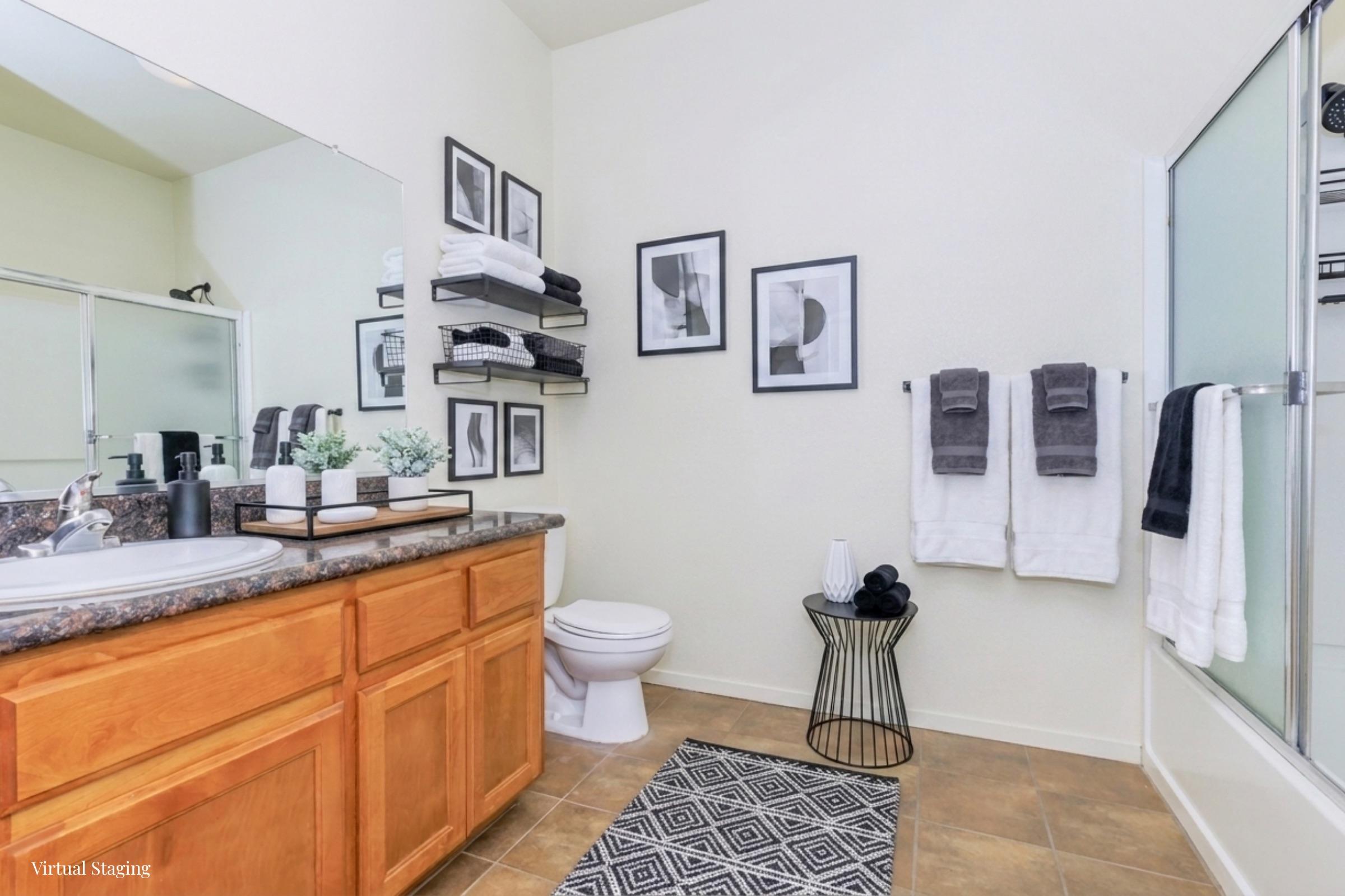 A modern bathroom featuring a wooden vanity with a large mirror, black and white framed artwork on the walls, and neatly arranged towels. A small black wire table holds decorative items. Neutral-colored tiles and a shower enclosure complete the clean, contemporary look.