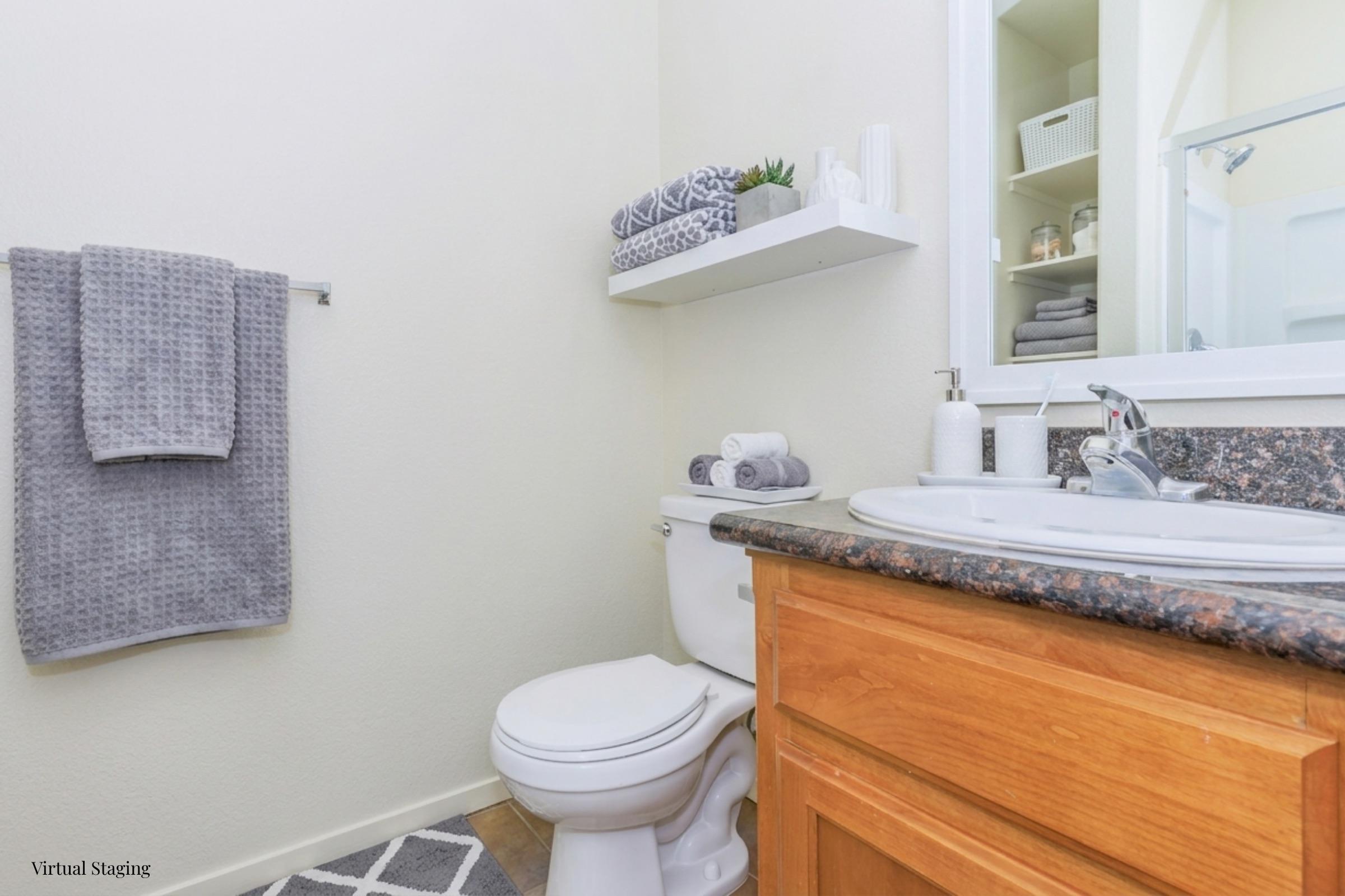 A clean and well-lit bathroom featuring a white toilet, a granite countertop with a sink, and shelves above with neatly folded towels. A gray towel hangs on a rack, and a decorative plant sits on the counter. The floor is covered with a stylish gray and white rug, adding a modern touch to the space.