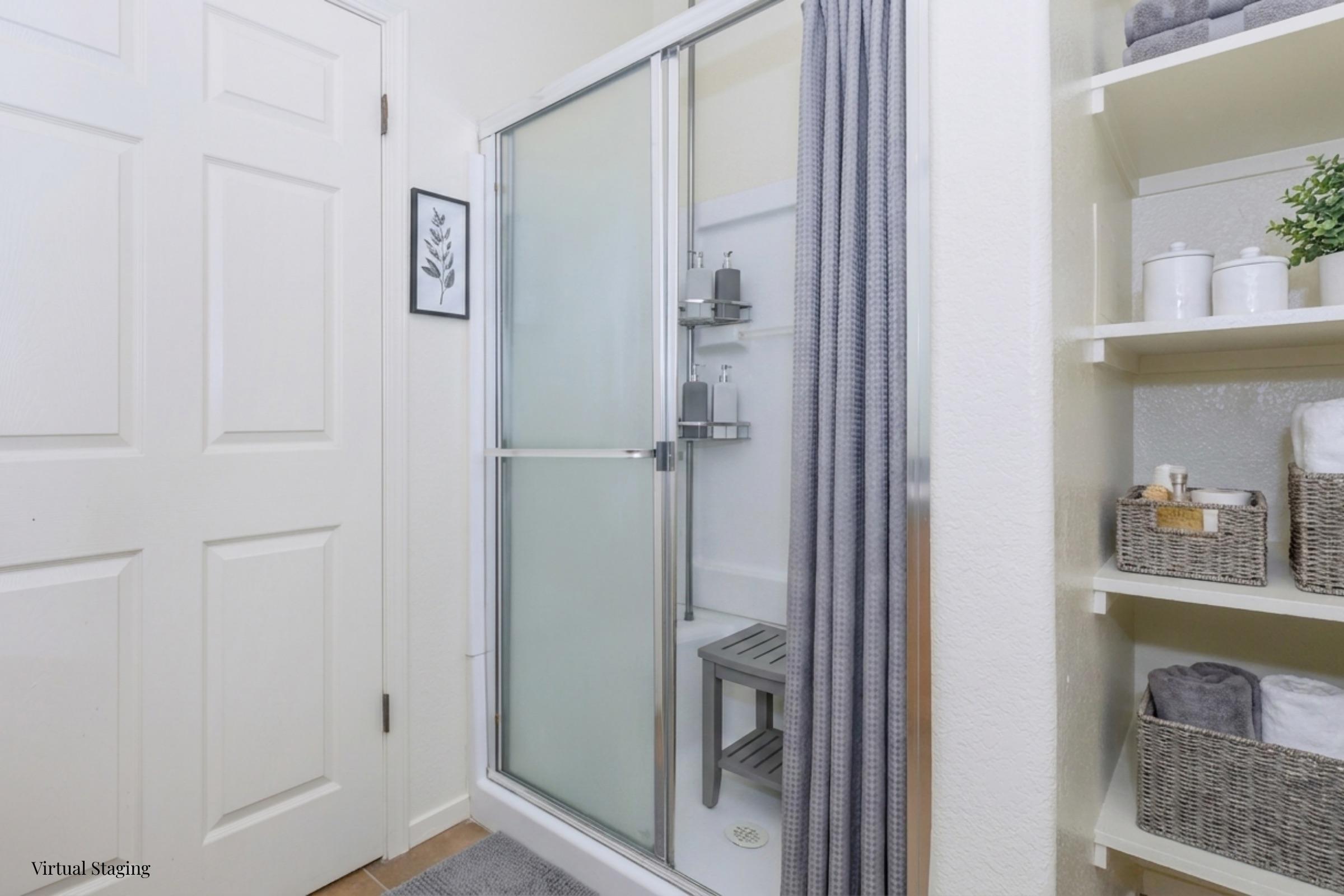 A clean and bright bathroom featuring a glass shower with a silver frame, a wooden shower stool, and neatly arranged toiletries. To the right, there are shelves with towel and storage baskets. A wall art piece adds a decorative touch, and the room is well-lit.