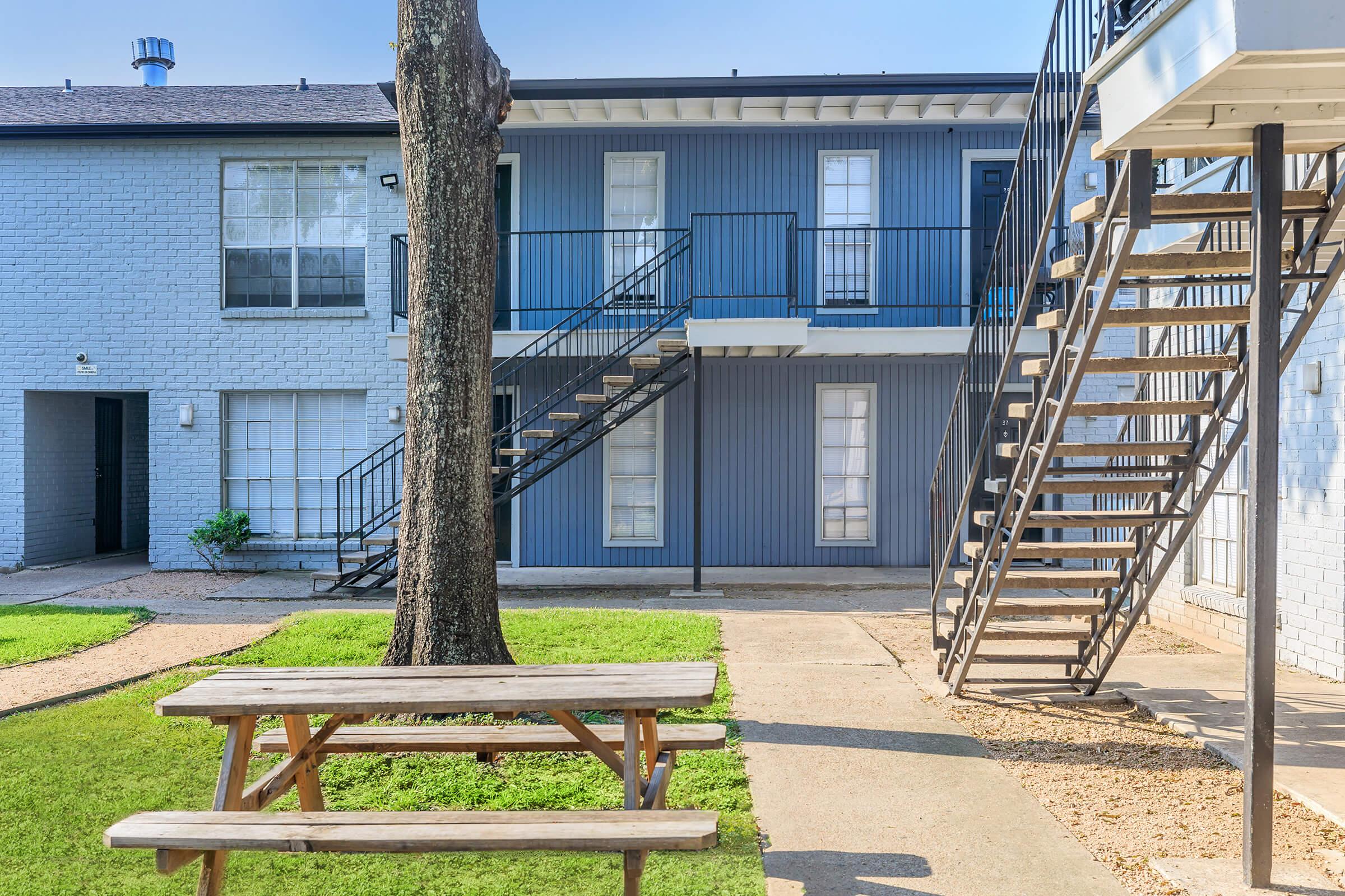 A courtyard view of a residential apartment complex featuring two buildings with staircases. In the foreground, there is a wooden picnic table on a grassy area, surrounded by pathways. The buildings are painted in light blue and white, and a tree is visible nearby.
