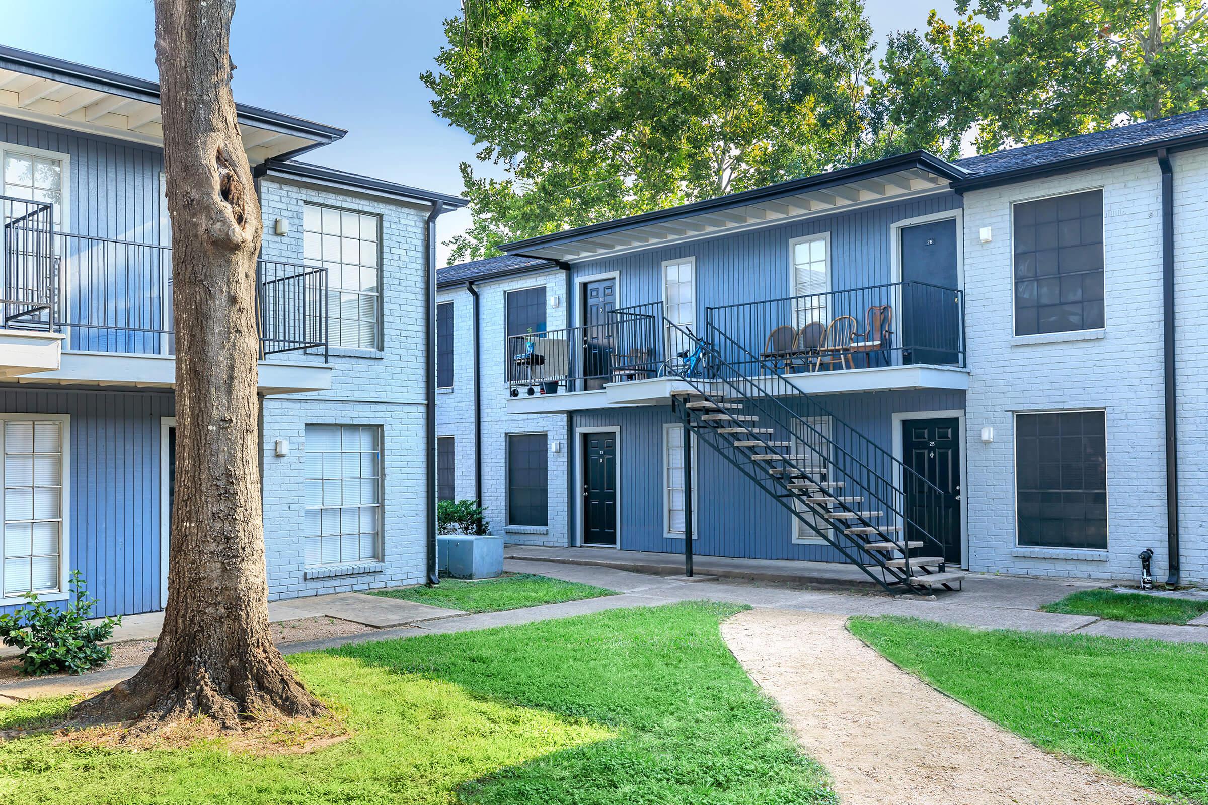 A view of a two-story apartment complex featuring light blue siding. The building has a central staircase with black railings, leading to balconies with chairs. Lush green grass surrounds the building, and a tree is visible in the foreground.