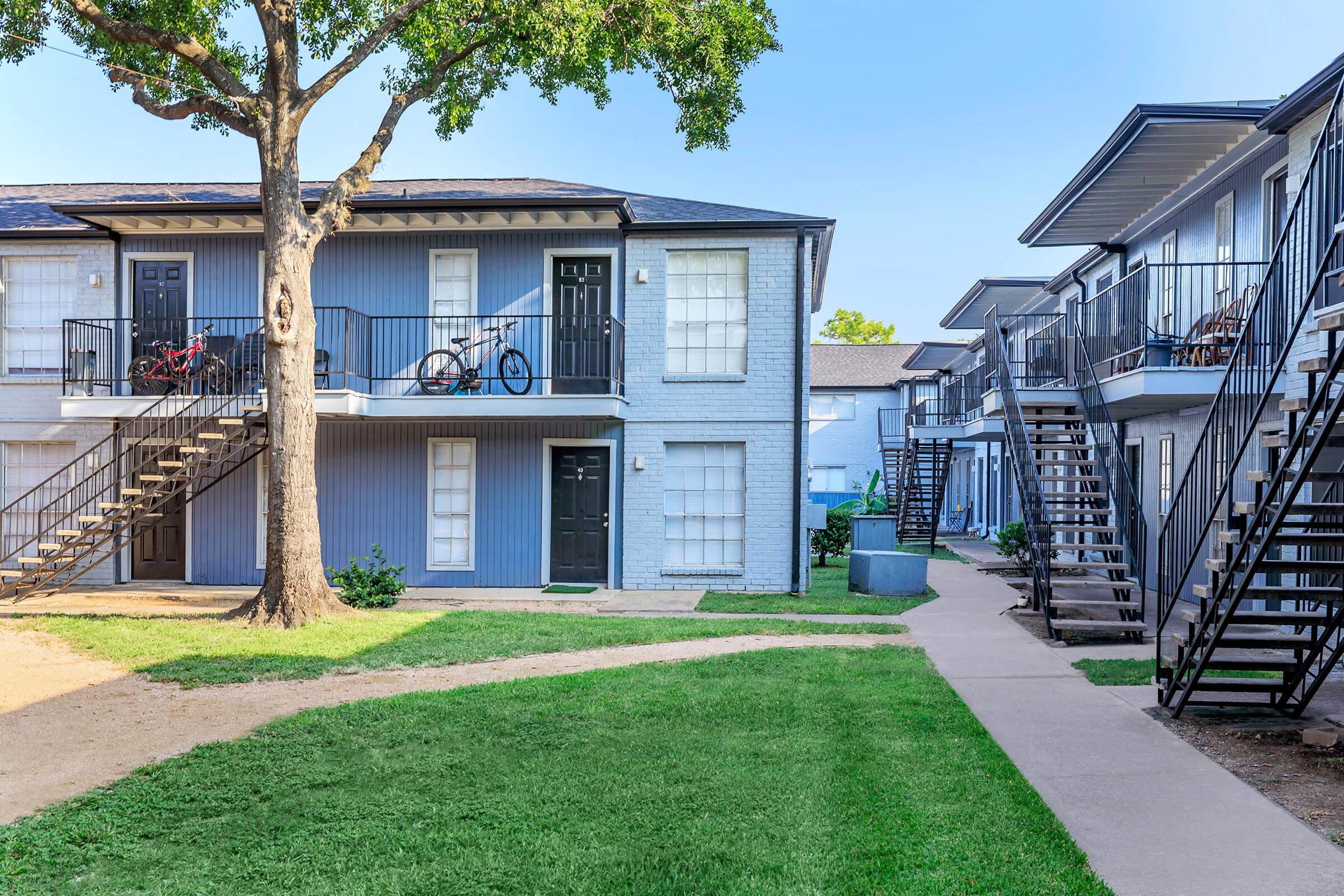 Two-story apartment complex surrounded by a grassy courtyard. The buildings feature stairs leading to upper units, with a tree and bike parked in front of one unit. Pathways connect the apartments, creating a welcoming outdoor space. Clear blue sky complements the scene.