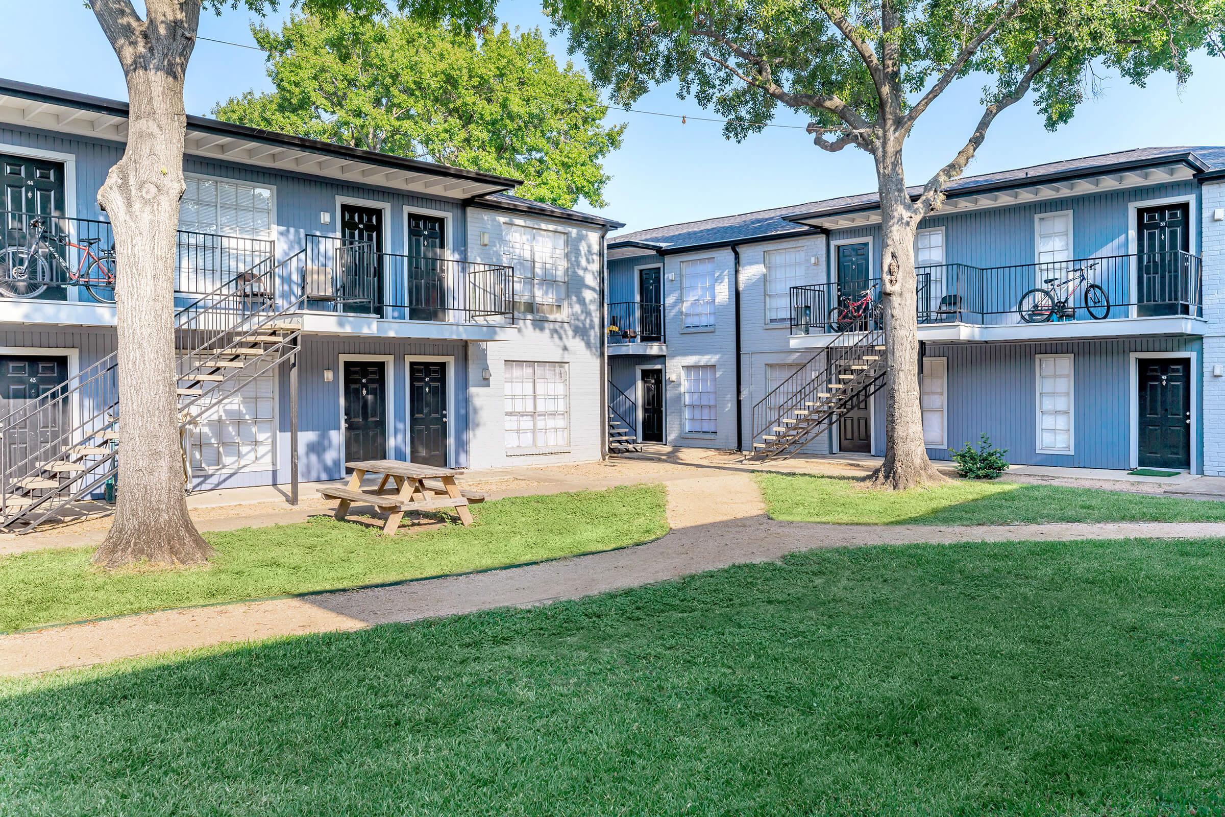 A view of a two-story apartment complex featuring multiple entrances, with a central grassy area and picnic table in the foreground. The buildings are painted in shades of blue and white, surrounded by trees. Pathways connect the apartments, enhancing the communal feel of the space.