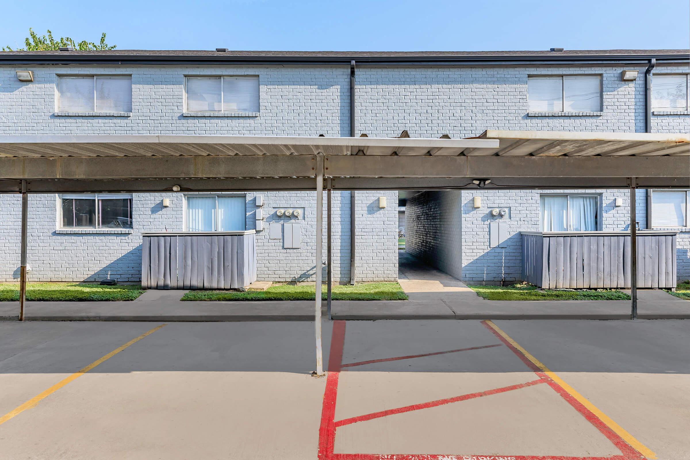 View of a residential building consisting of two-story units, featuring large windows and outdoor access. The foreground includes a shaded parking area with a visible red line marking. The overall setting appears bright and well-maintained, with green grass lining the walkway.
