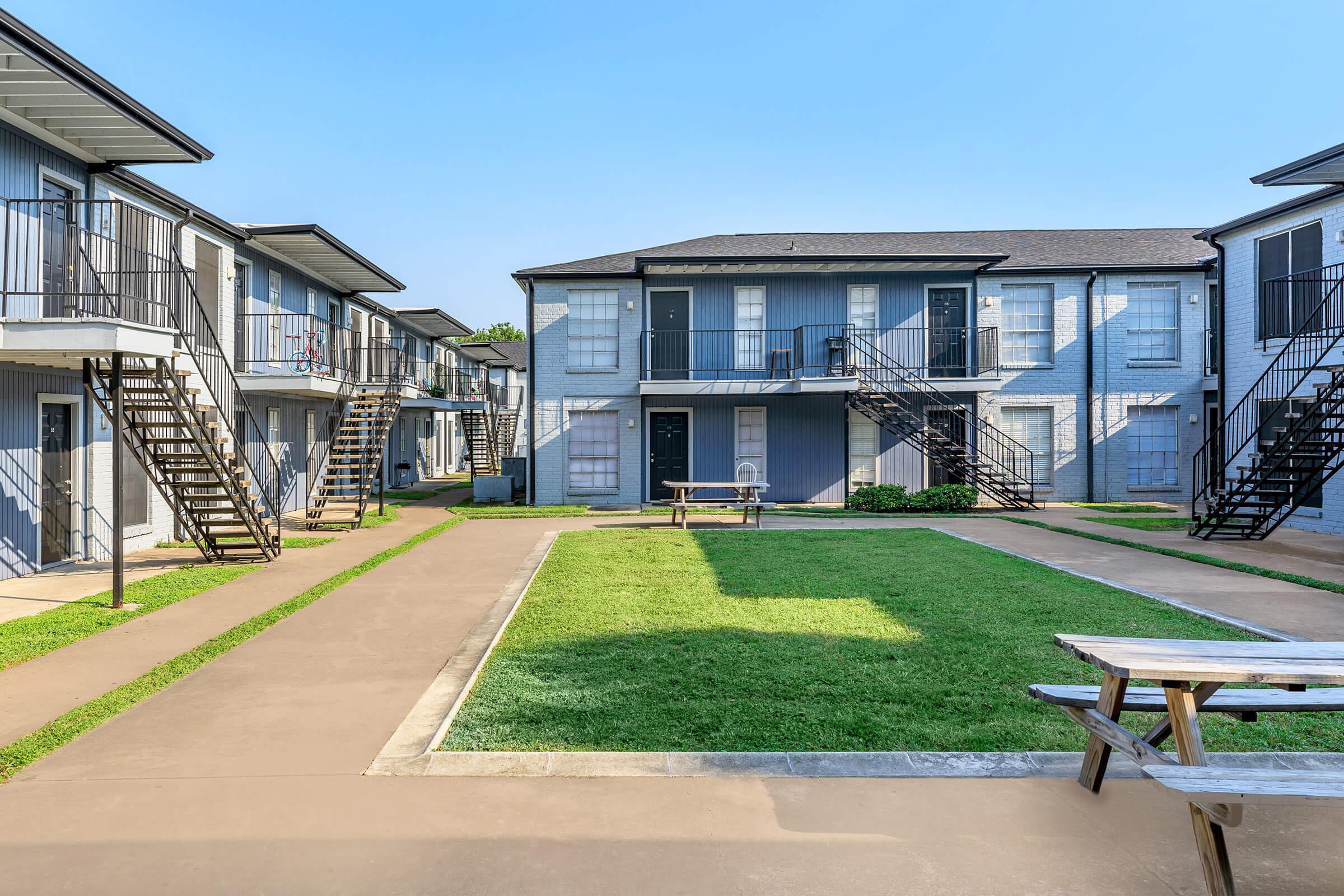 A view of an apartment complex featuring two-story buildings with balconies and outdoor staircases. The central area has a well-maintained grassy lawn and a small picnic table. The sky is clear and blue, adding to the inviting atmosphere of the property.