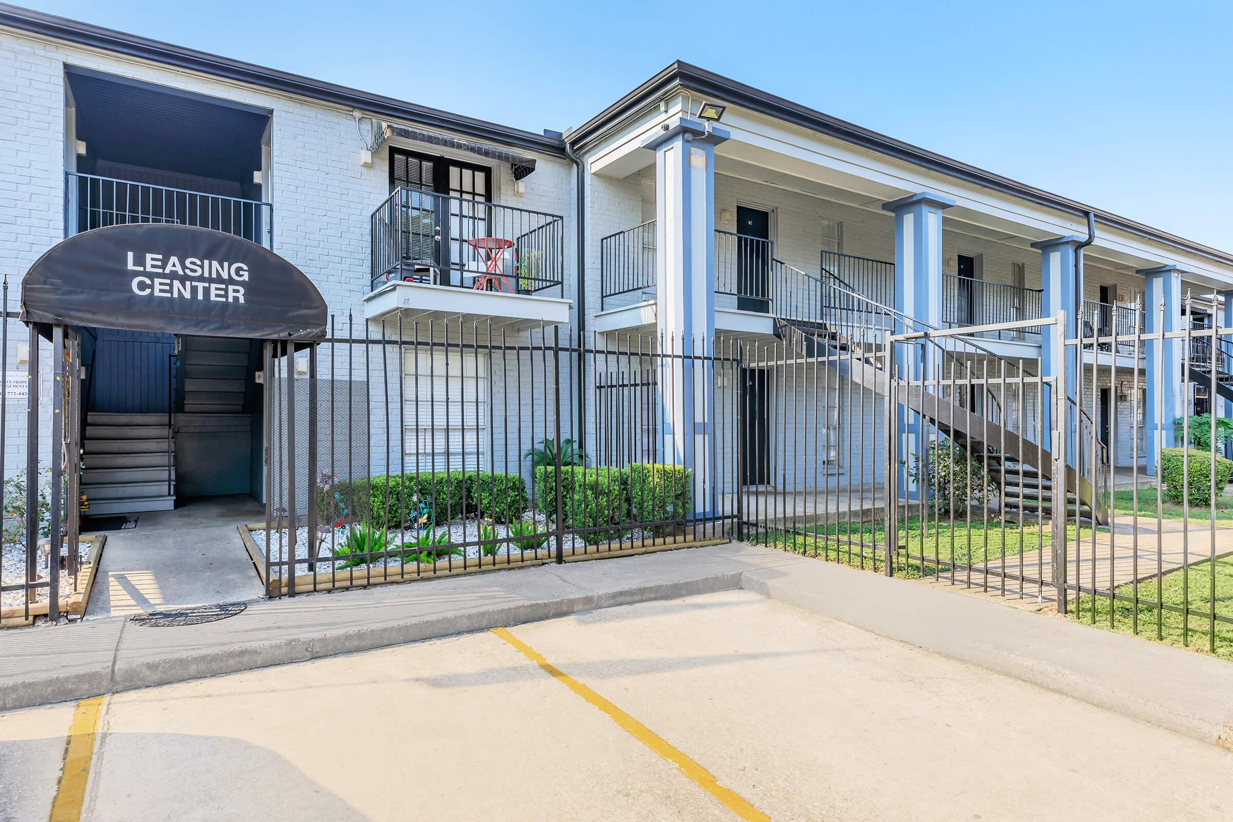 Exterior view of a leasing center for an apartment complex, featuring a fenced area, two-story building with balconies, and a parking lot. The entrance is marked with a canopy sign reading "Leasing Center." Lush greenery is visible along the path and near the entrance. Clear blue sky in the background.