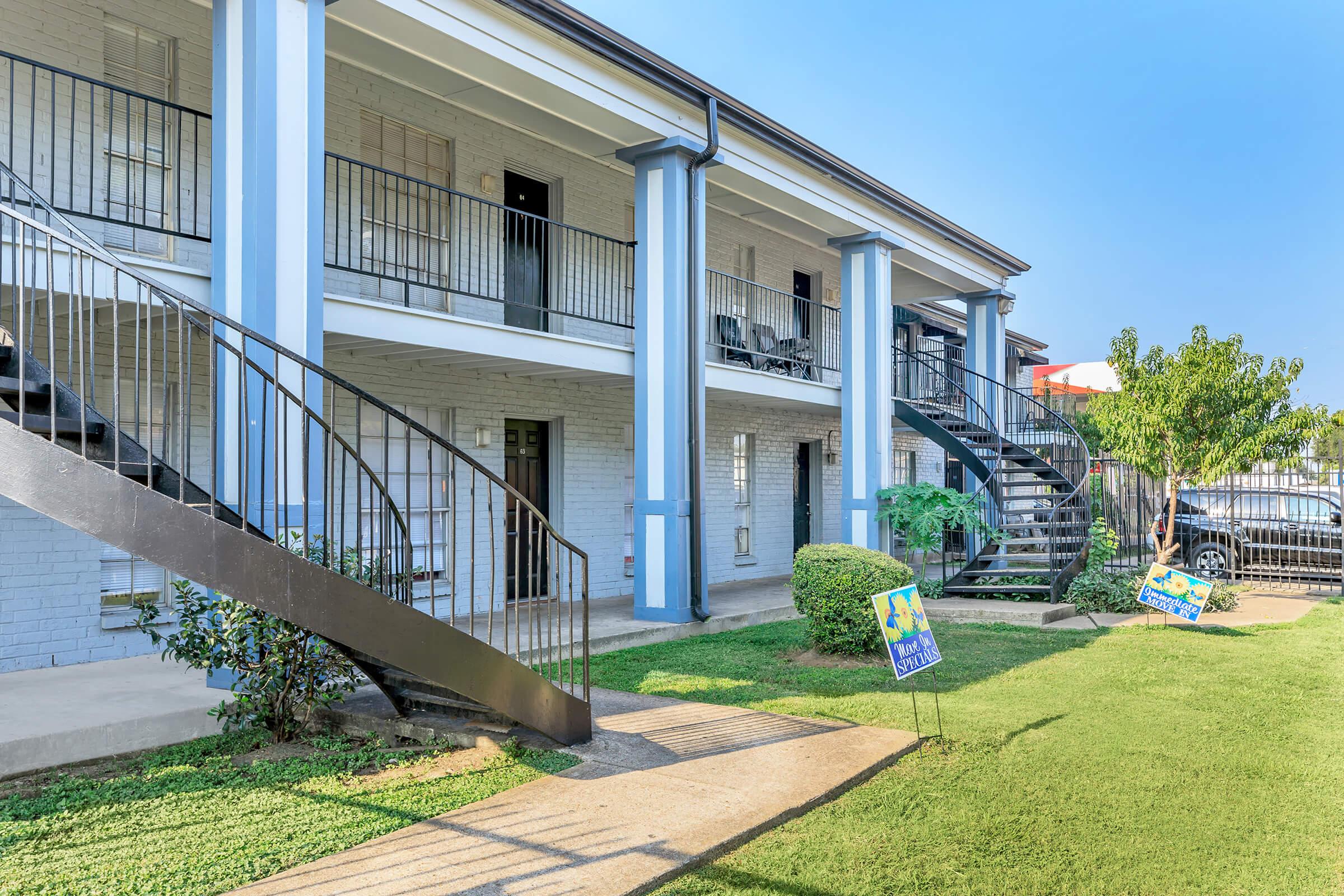 View of an apartment building with two levels. The exterior features a light brick façade and blue pillars. A winding staircase leads to the second floor. Lush green grass and small shrubs line the path, with signs advertising apartments visible in the foreground. Clear blue sky overhead.