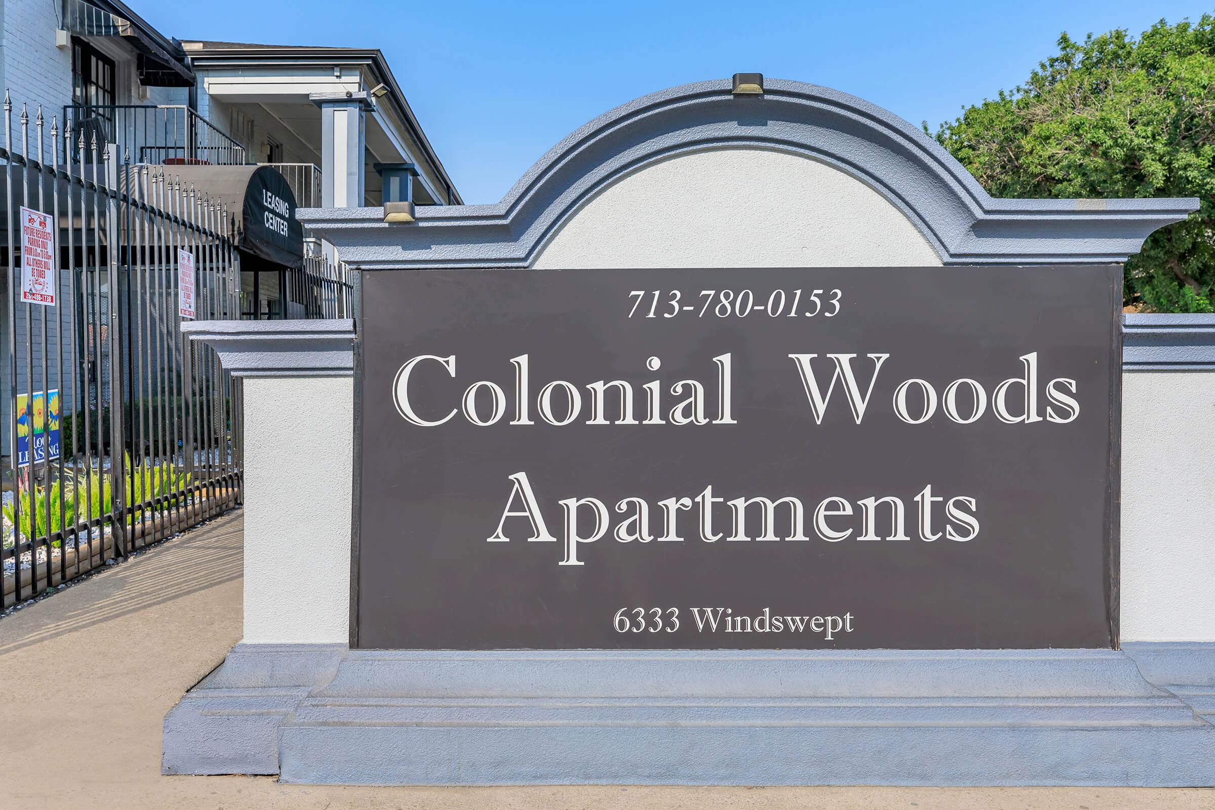 Sign for Colonial Woods Apartments, displaying the address "6333 Windswept" and a phone number "713-780-0153." The entrance features a decorative archway, and the surrounding area is well-maintained with greenery visible.