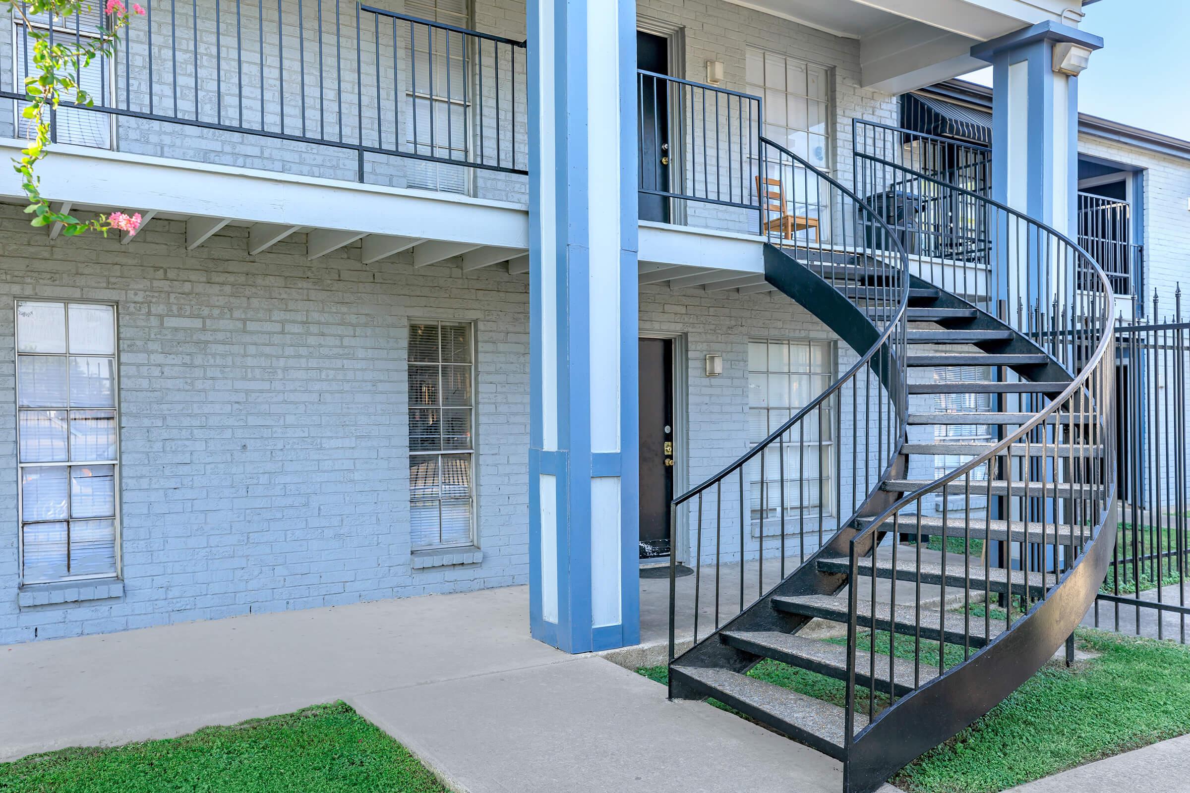 A modern building with a light gray brick exterior featuring a spiral staircase leading to the second floor. The ground level has a small concrete patio and two windows with white frames. The area is landscaped with green grass and a flowering vine near the staircase.