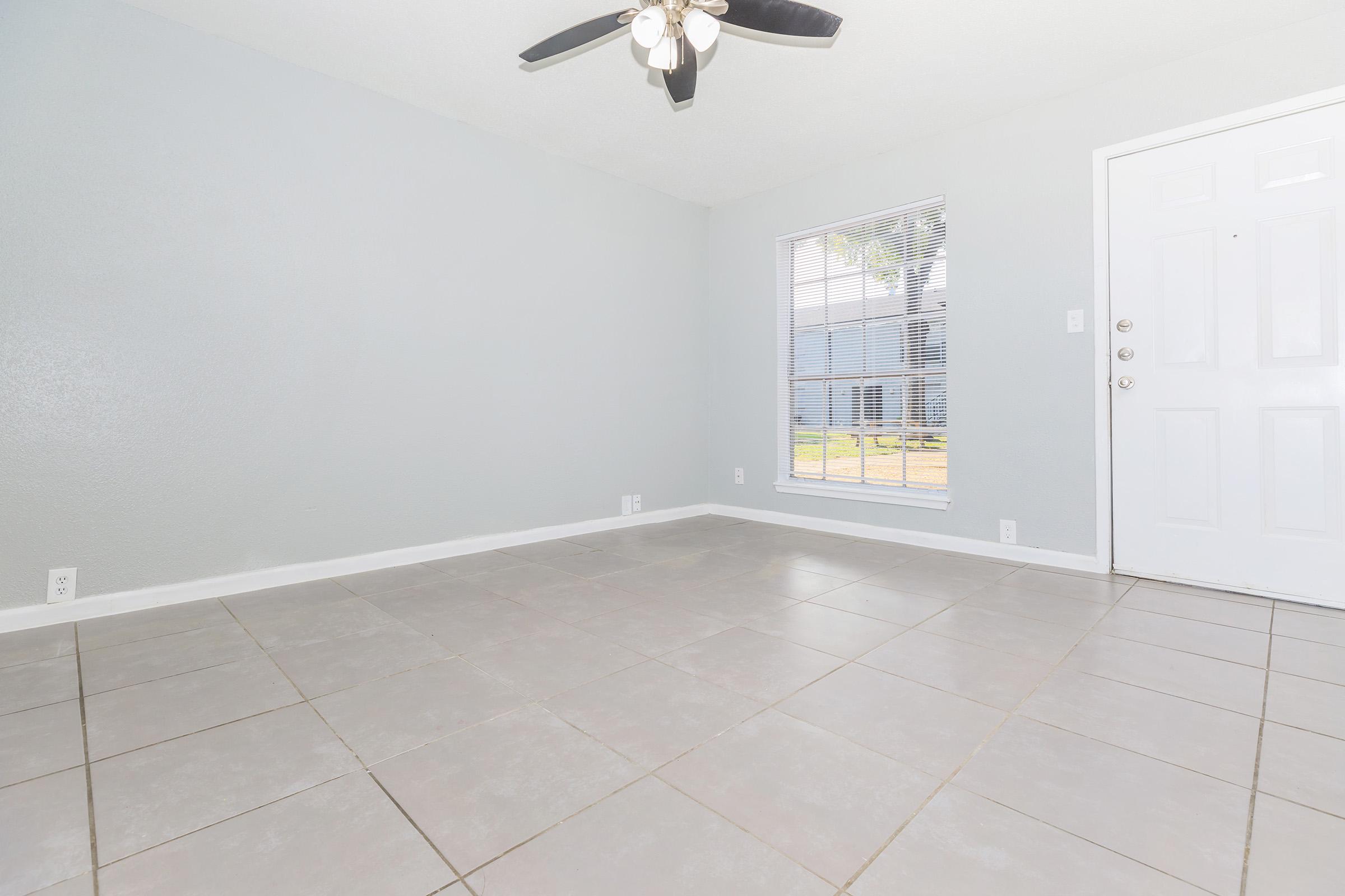 An empty room with gray walls and tiled flooring. A ceiling fan is visible above, and there's a large window letting in natural light. The door on the right leads outside. The space appears clean and unoccupied, making it suitable for various uses or furnishings.