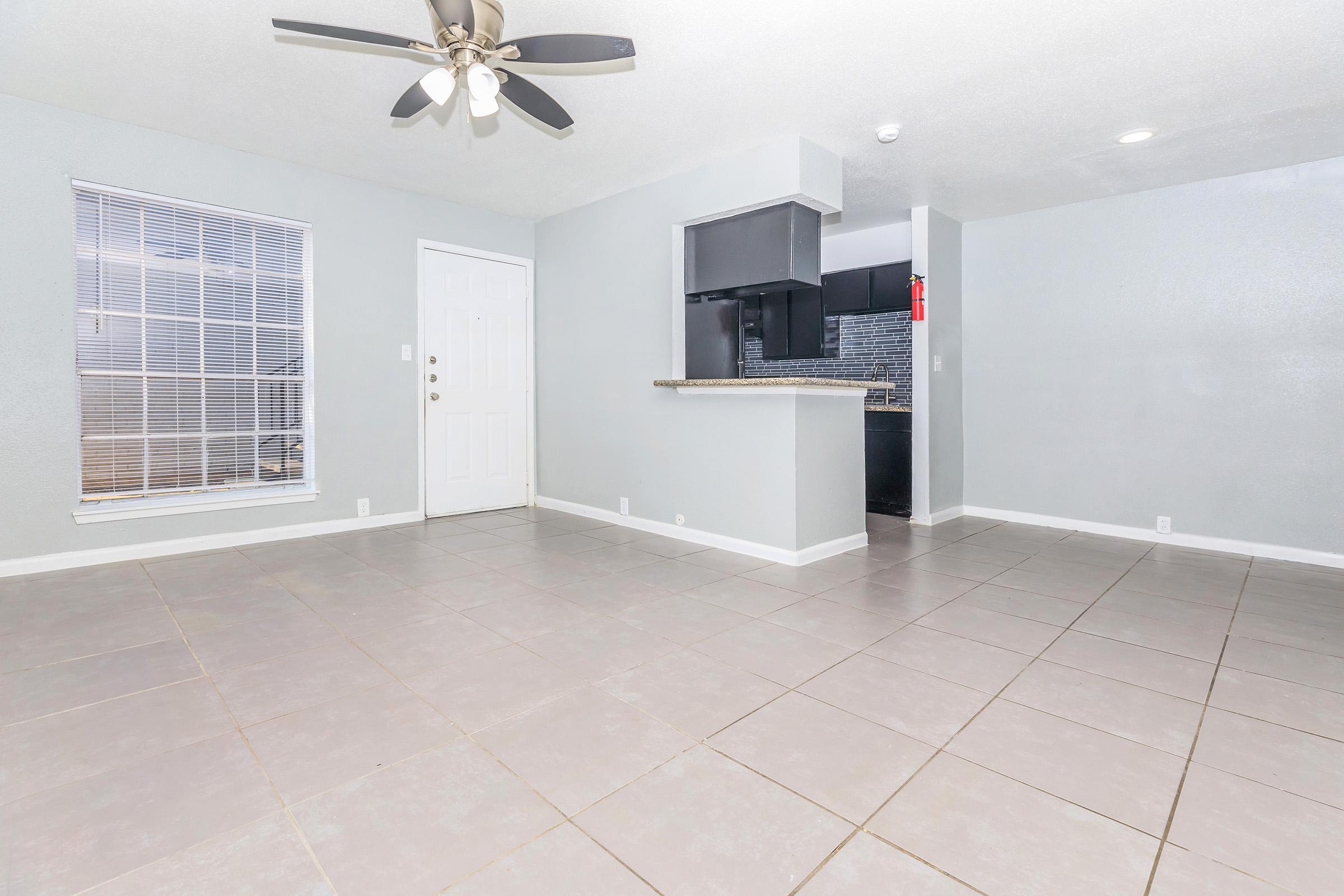 Spacious interior of an empty room with tiled flooring, a ceiling fan, and a door leading outside. A kitchen area is visible in the background with dark cabinets and a countertop, and a large window allows natural light to enter the space. The walls are painted light gray.