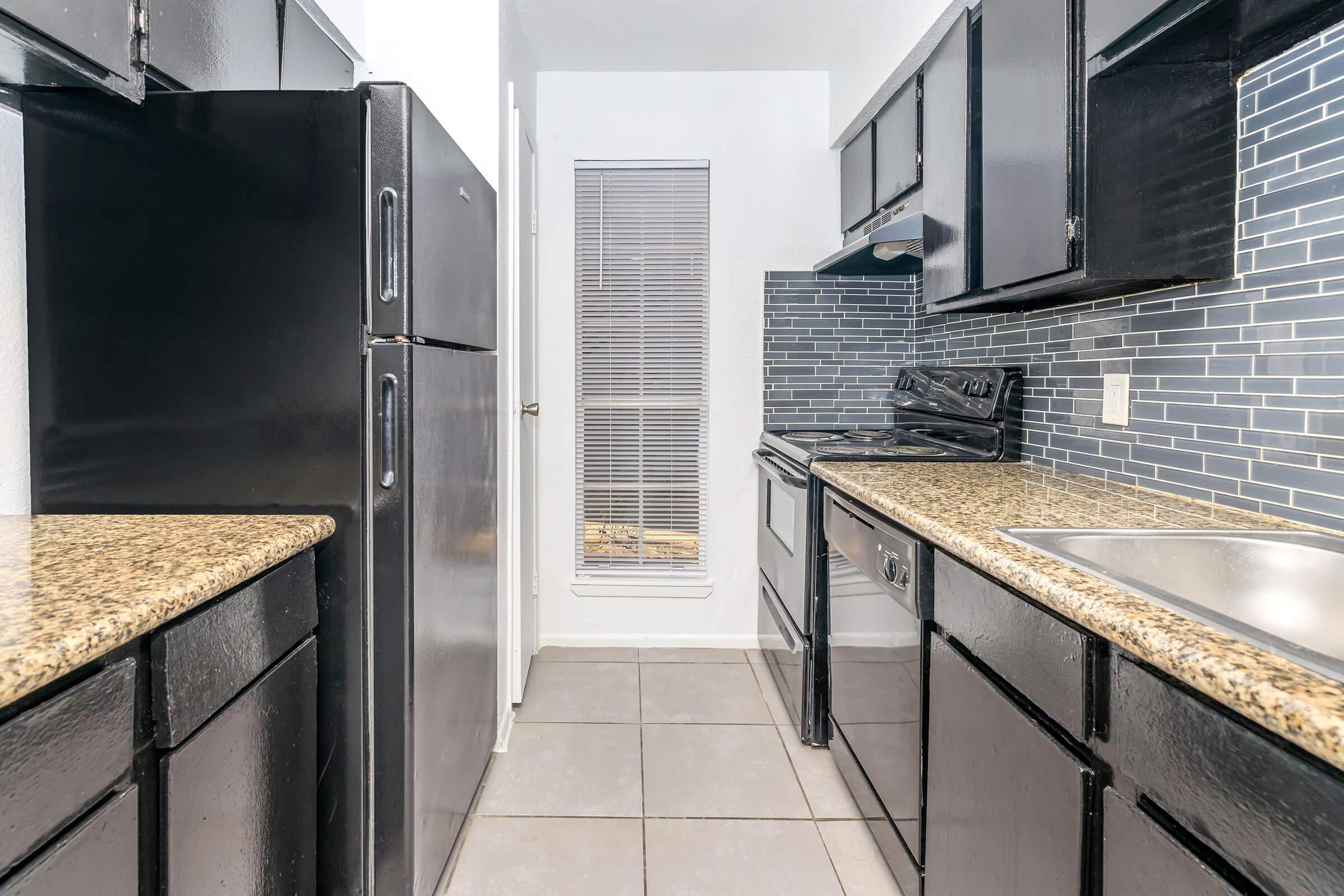 A modern kitchen featuring black appliances, including a refrigerator and stove, along with dark cabinets and a granite countertop. The backsplash has mosaic tiles in blue tones, and there is a window with blinds providing natural light. The flooring is tiled.