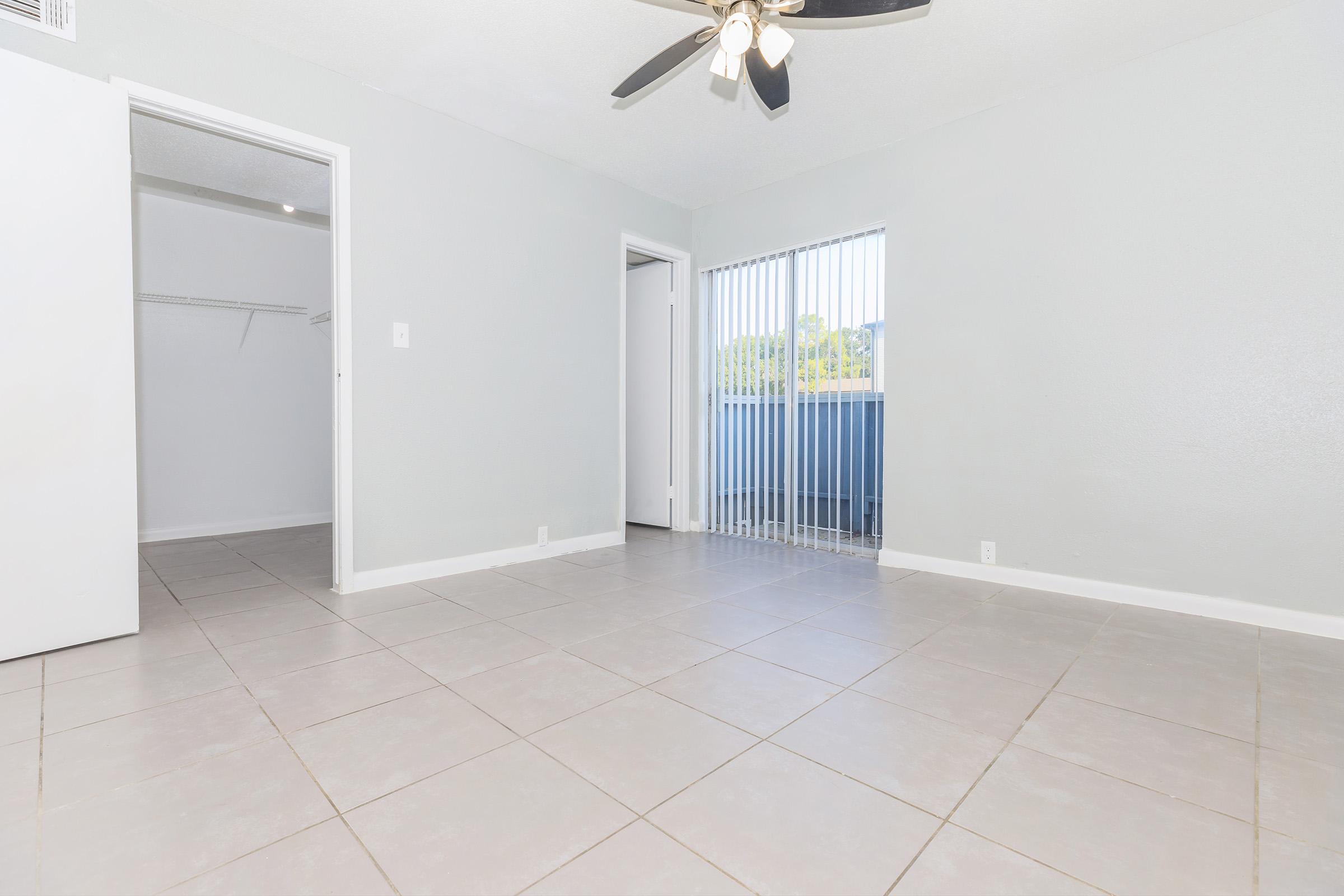 A spacious, light-filled room with gray walls and tiled flooring. It features a ceiling fan and large sliding doors with vertical blinds leading to a balcony. An open closet space is visible on the left, and another door is seen on the right, suggesting access to another area.
