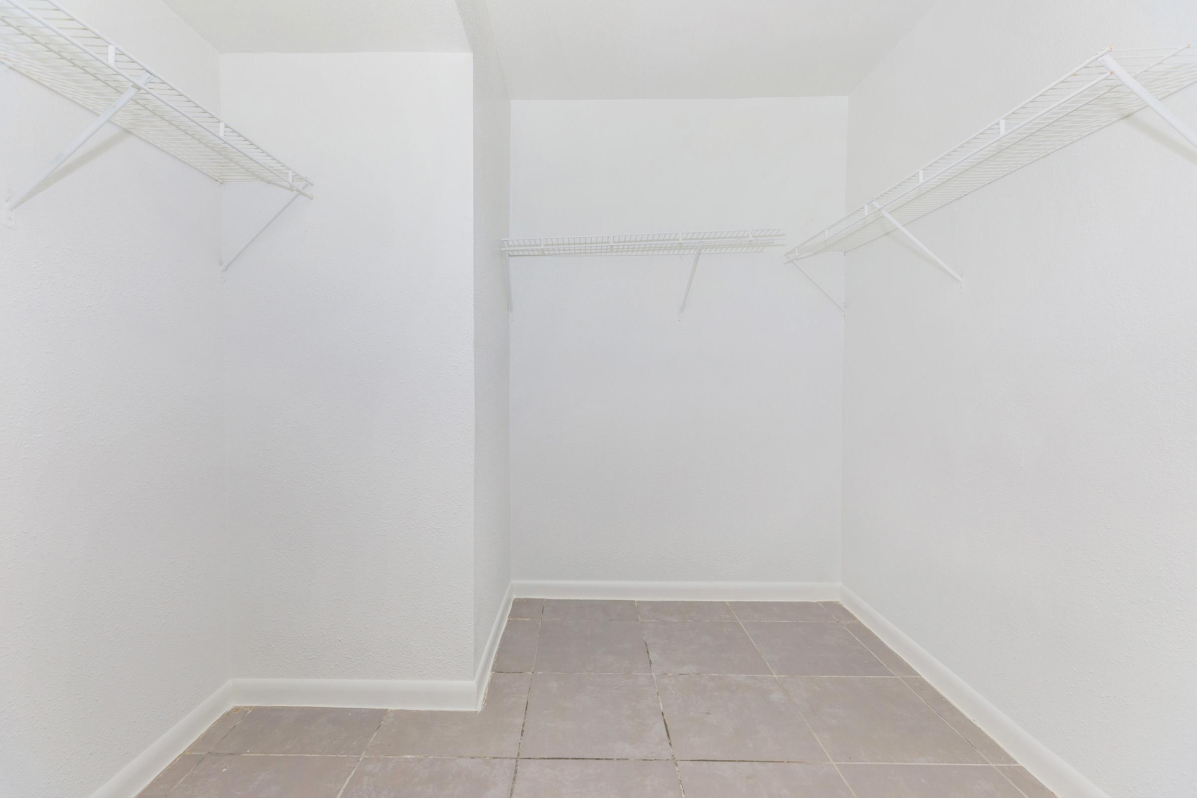 A spacious, empty closet featuring white walls and gray tile flooring. There are wire shelves mounted on two walls, providing storage options. The room is well-lit, creating an airy feel, and lacks any furniture or personal items.