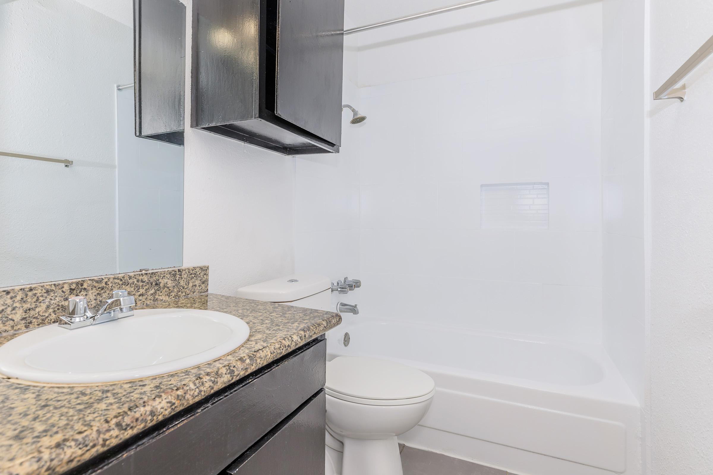 A clean bathroom featuring a white bathtub and shower combination, a granite countertop sink with a faucet, and dark wooden cabinets. The walls are painted white, and there is a mirror above the sink. Bright lighting enhances the space, giving it a fresh and modern look.