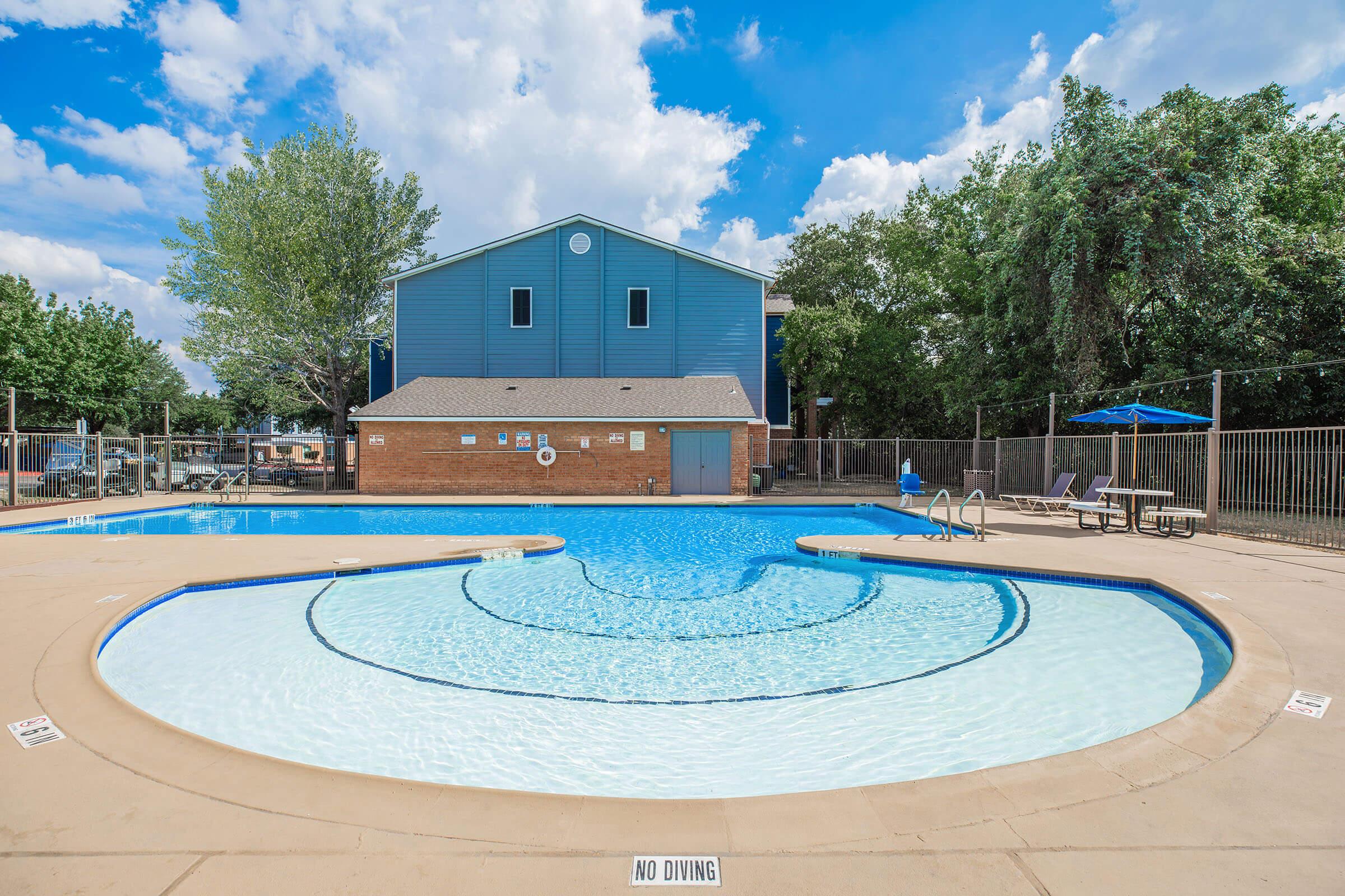 A swimming pool with a curved design, surrounded by a fence and lounge chairs. In the background, there is a two-story blue building and green trees. The sky is bright with clouds, and a "No Diving" sign is visible near the pool's edge.