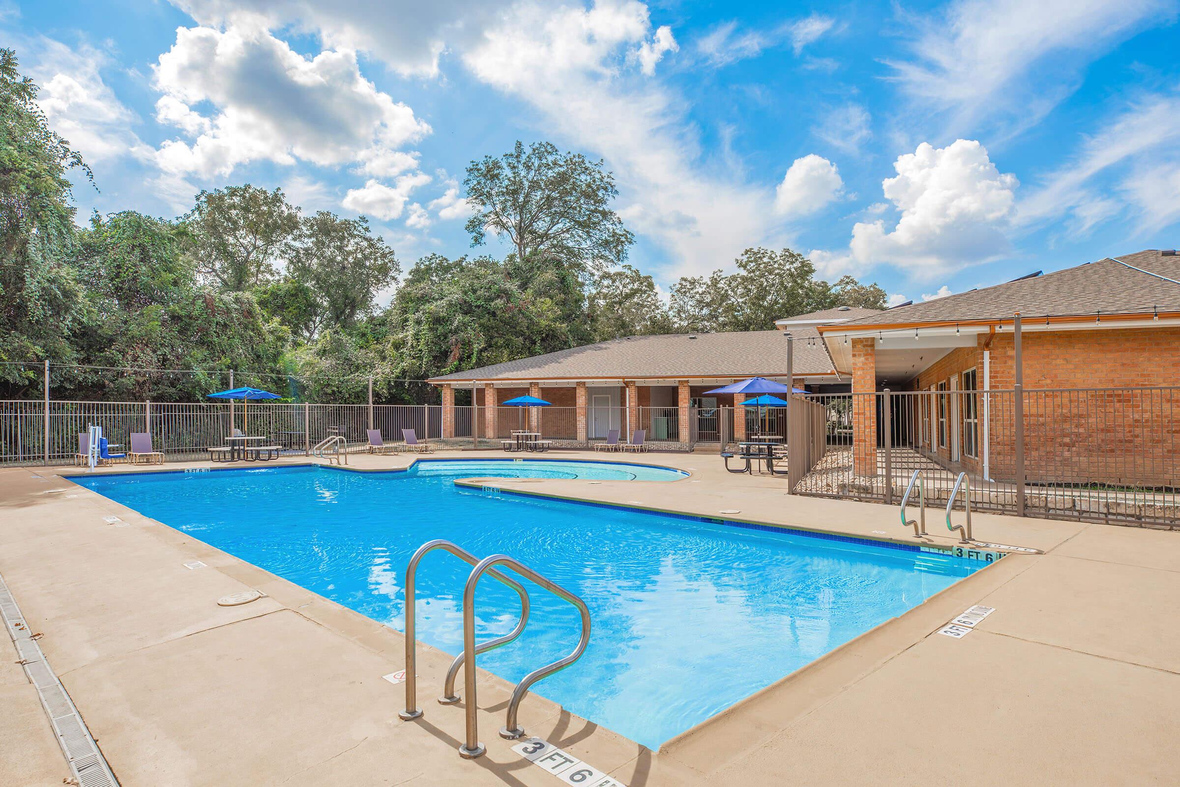 A clear blue swimming pool surrounded by a tan concrete deck. There are lounge chairs and umbrellas on one side, while a brick building with several windows is visible in the background. Lush green trees provide shade and a bright sky with fluffy clouds is overhead.