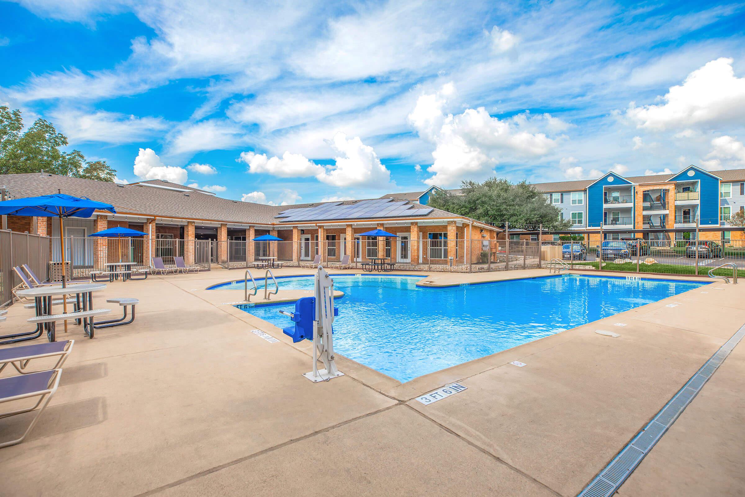 A clear swimming pool surrounded by lounge chairs and umbrellas, with a blue sky and fluffy clouds above. In the background, there are apartment buildings with a mix of colors. The area is well-maintained, creating a relaxing outdoor environment.