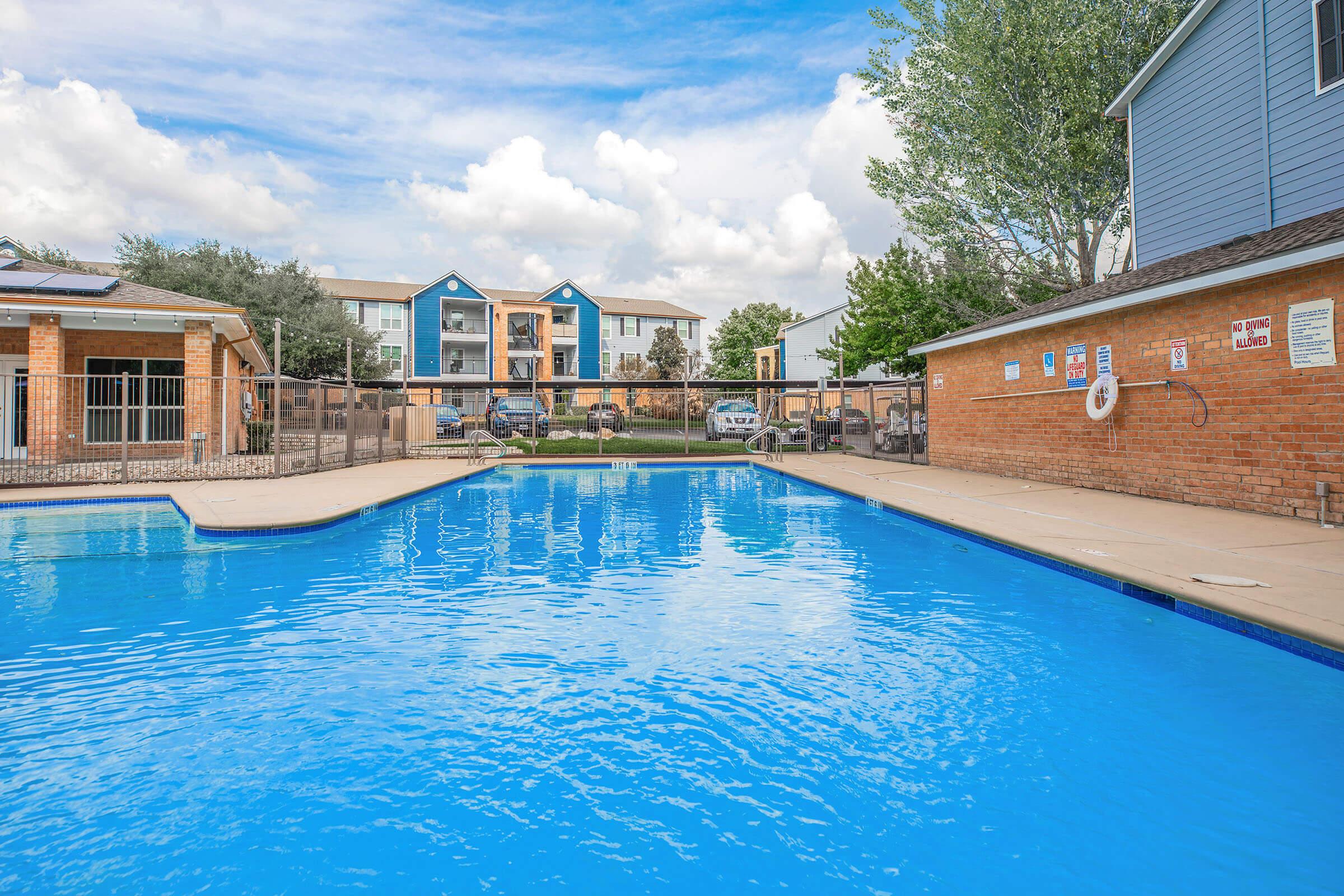 A clear blue swimming pool surrounded by a patio, with lounge chairs and trees in the background. An apartment complex with colorful exterior is visible across from the pool area, under a partly cloudy sky.