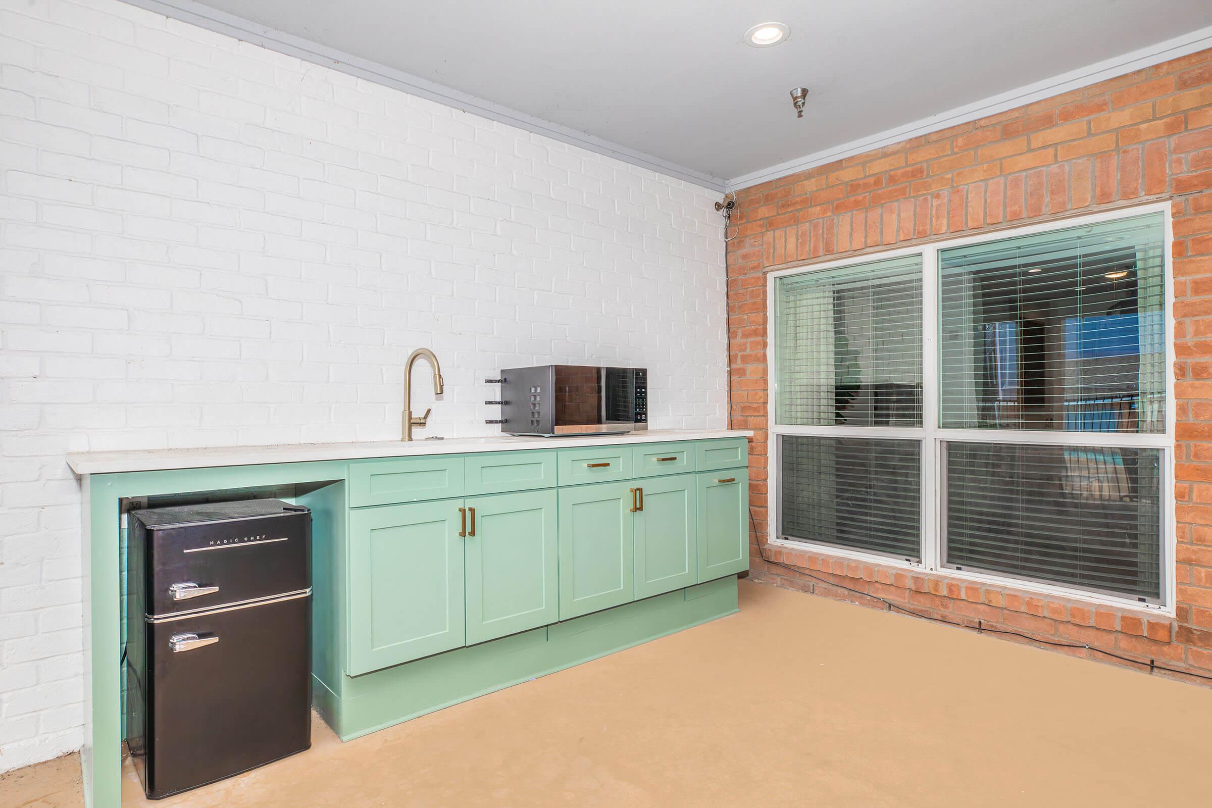 A corner of a kitchen featuring a light green cabinet with gold handles, a black microwave on top, and a black mini fridge beneath. The wall has a white brick finish, and there's a large window with a view, allowing natural light to brighten the space.